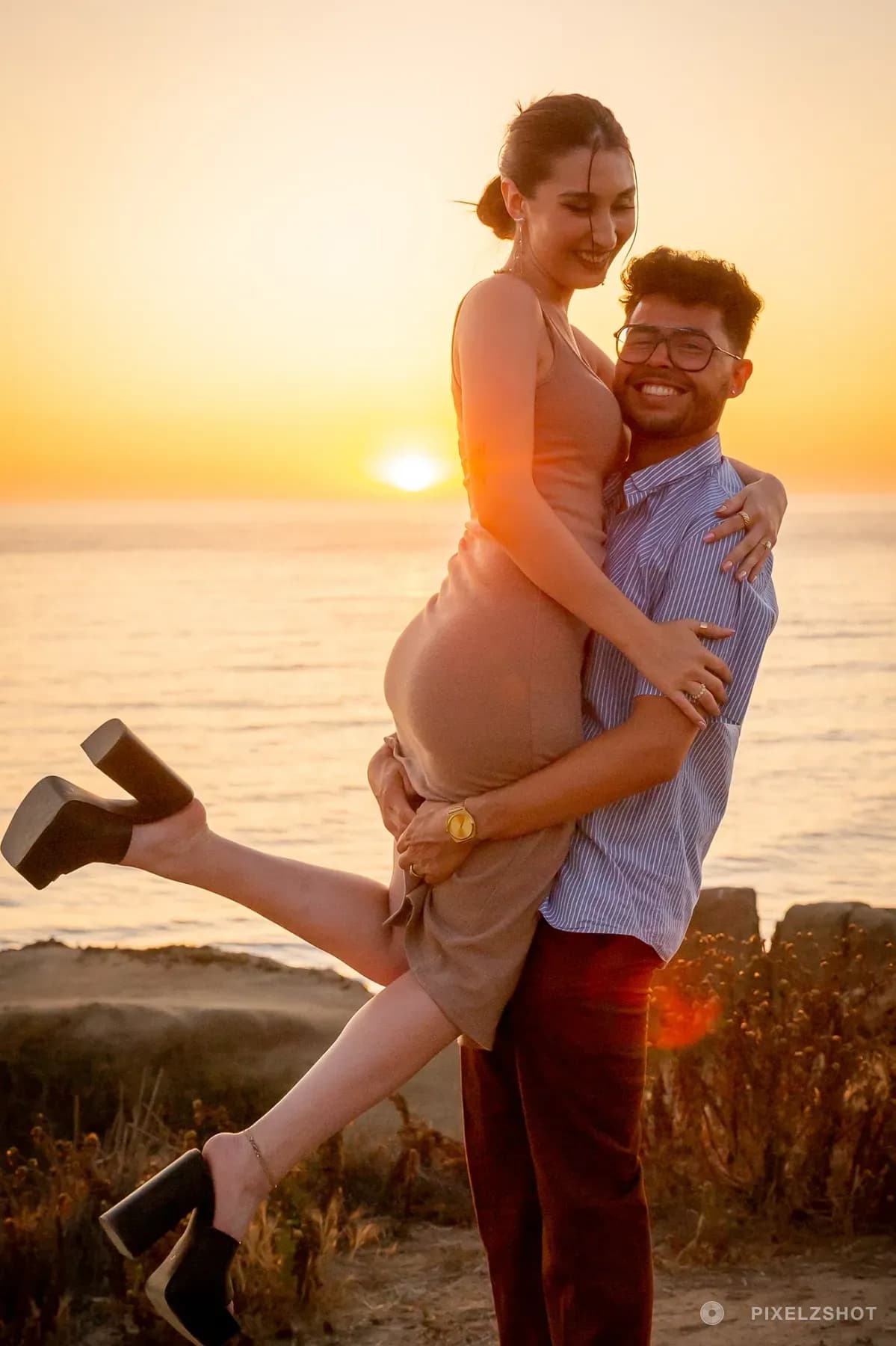 Danielle and Mathew laughing as one lifts the other off her feet, the sun dropping to the horizon behind them