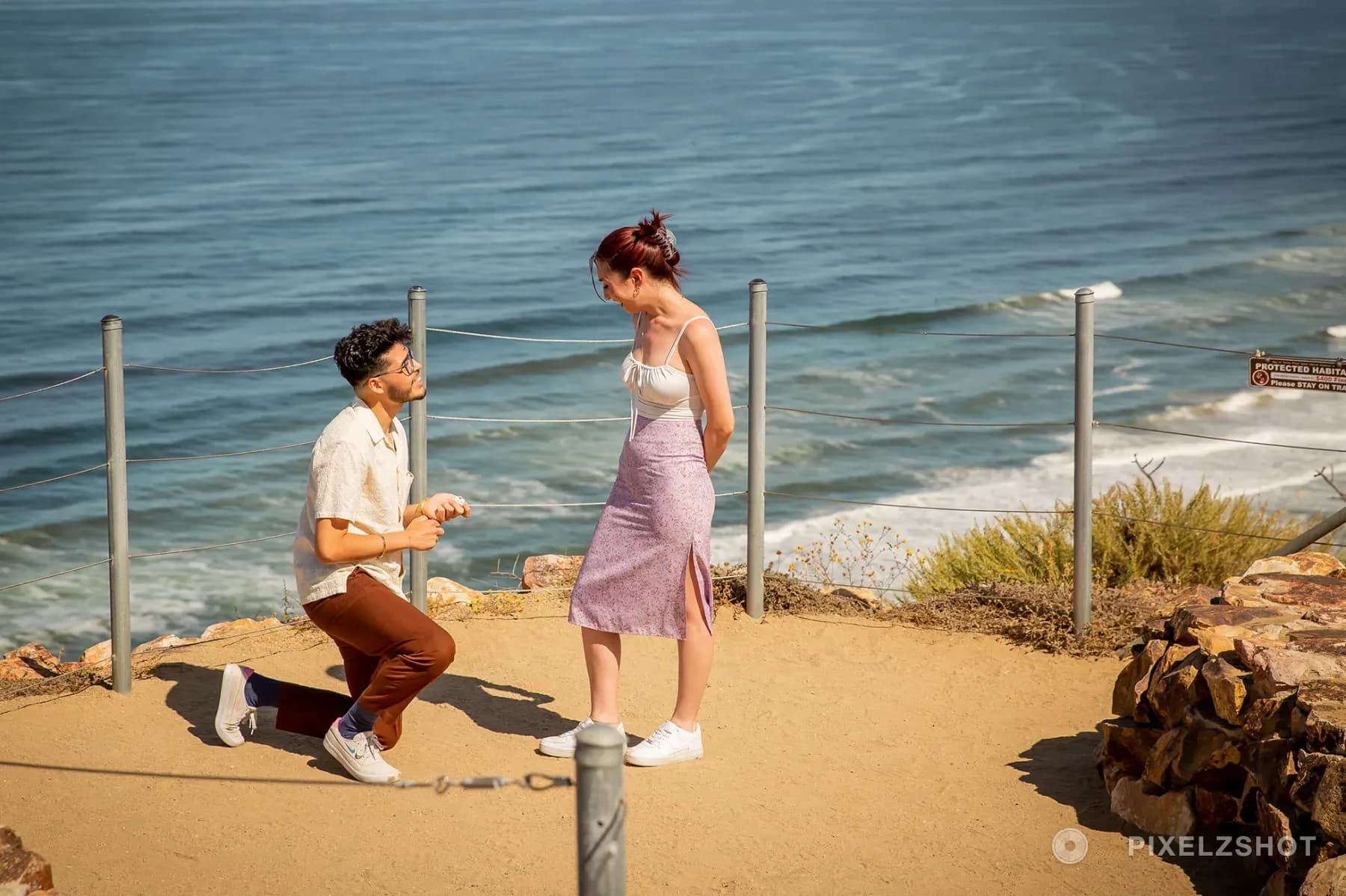 The moment of the proposal at a clifftop overlook, one of them down on one knee holding out the ring above the Pacific