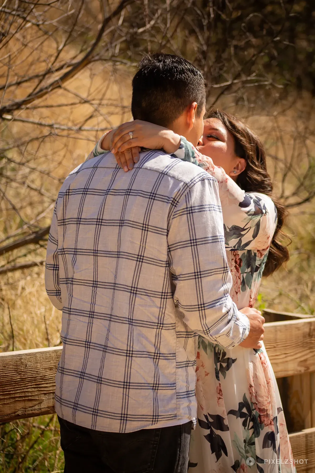 The couple in a close embrace beside a split-rail fence, one hand and its engagement ring resting on a shoulder