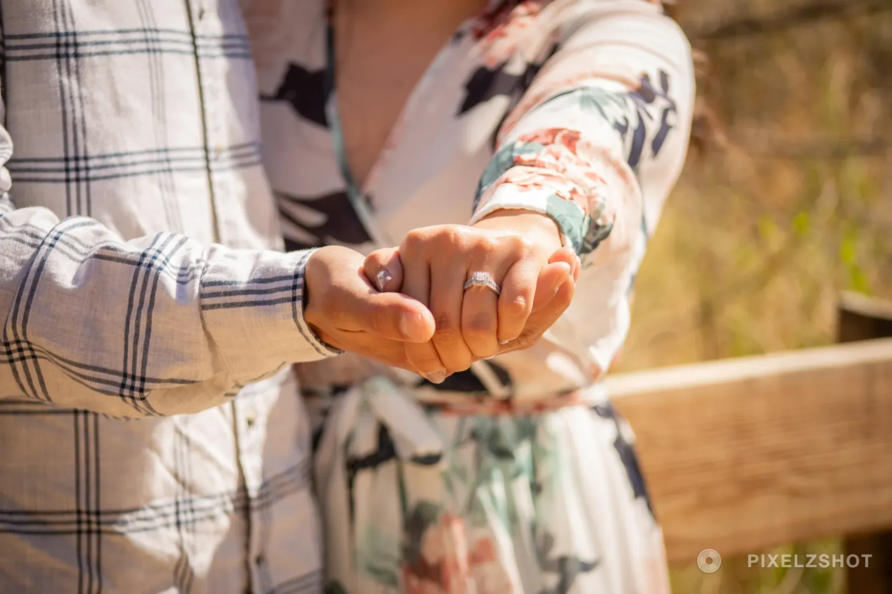 Their joined hands held out toward the camera with the engagement ring in focus and the trail fence soft behind