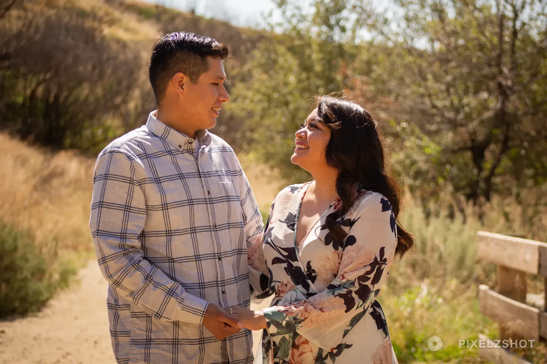 The couple standing hand in hand on a sunlit dirt trail, turned toward each other mid-laugh