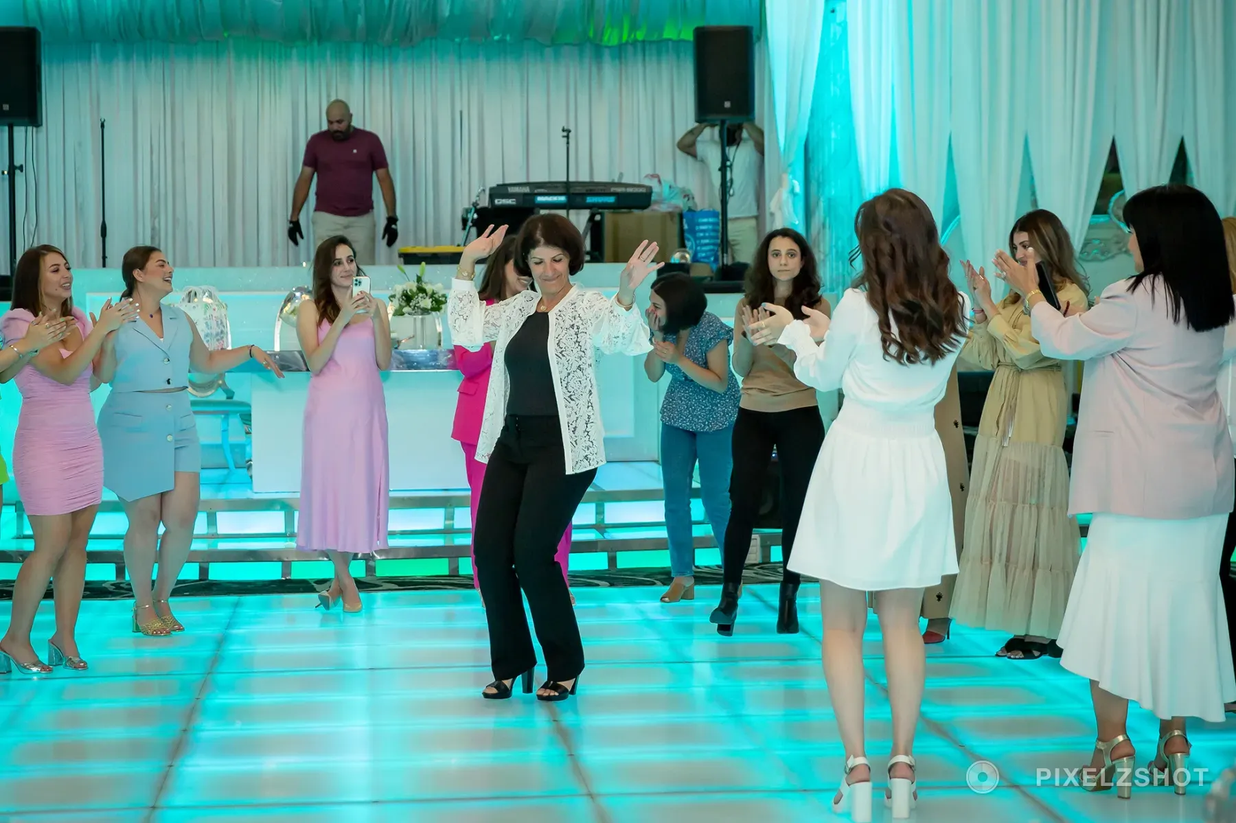 A guest dancing with both arms up in the centre of the floor, the circle around her clapping and filming