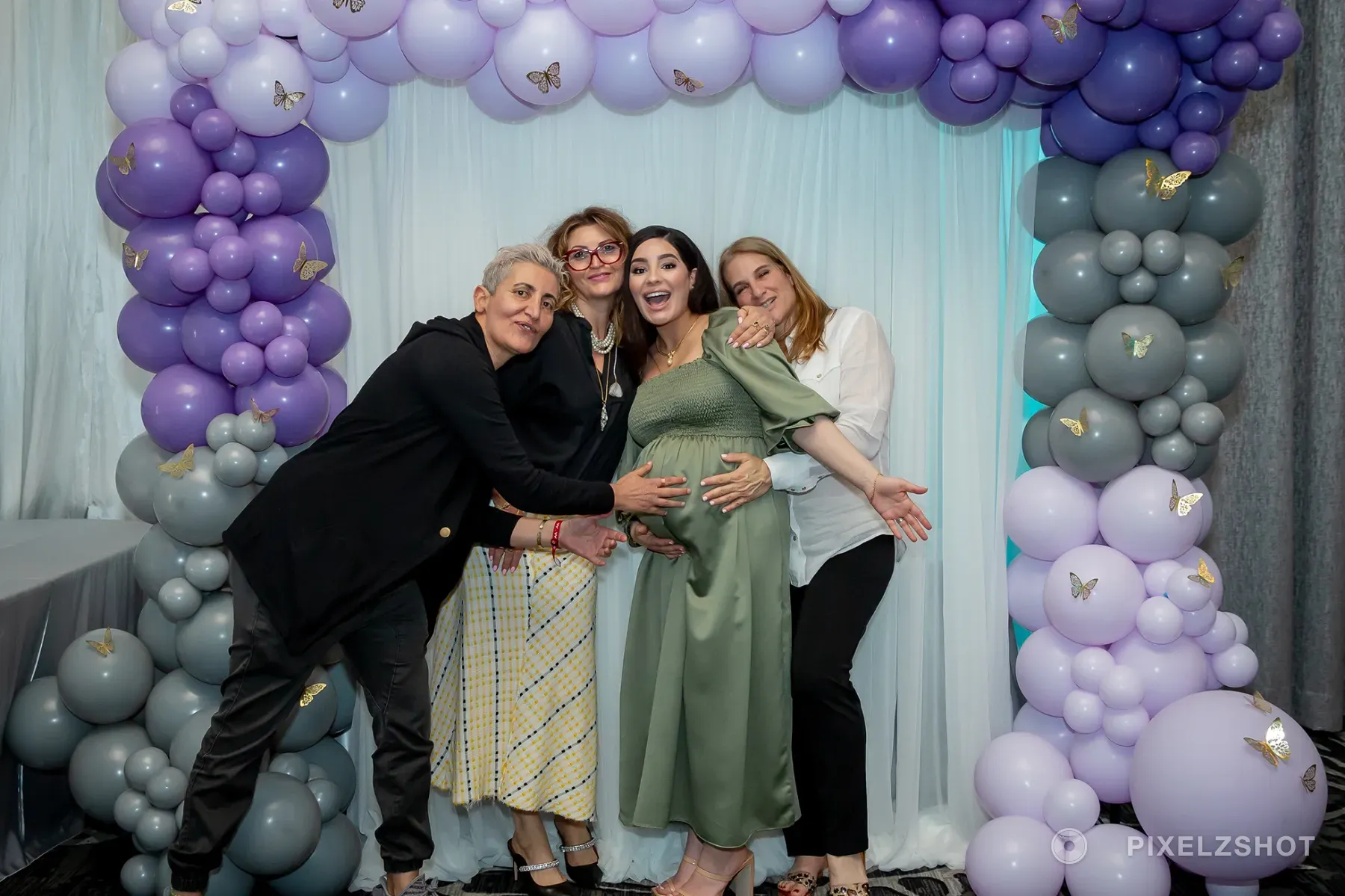 Alia laughing in a sage green dress while three guests lean in around her, every hand on the bump, under a lilac balloon arch