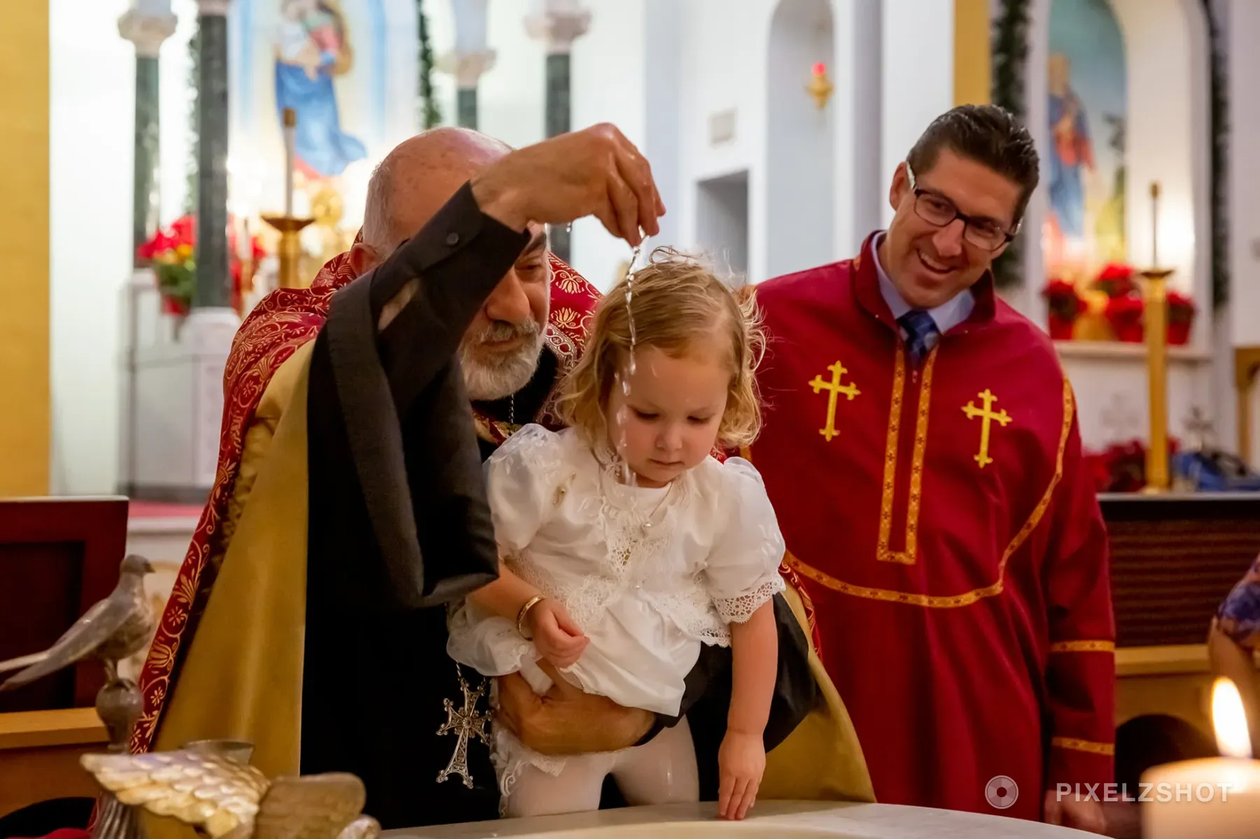 The priest pouring water over Sierra's head at the font, a deacon in red vestments smiling alongside