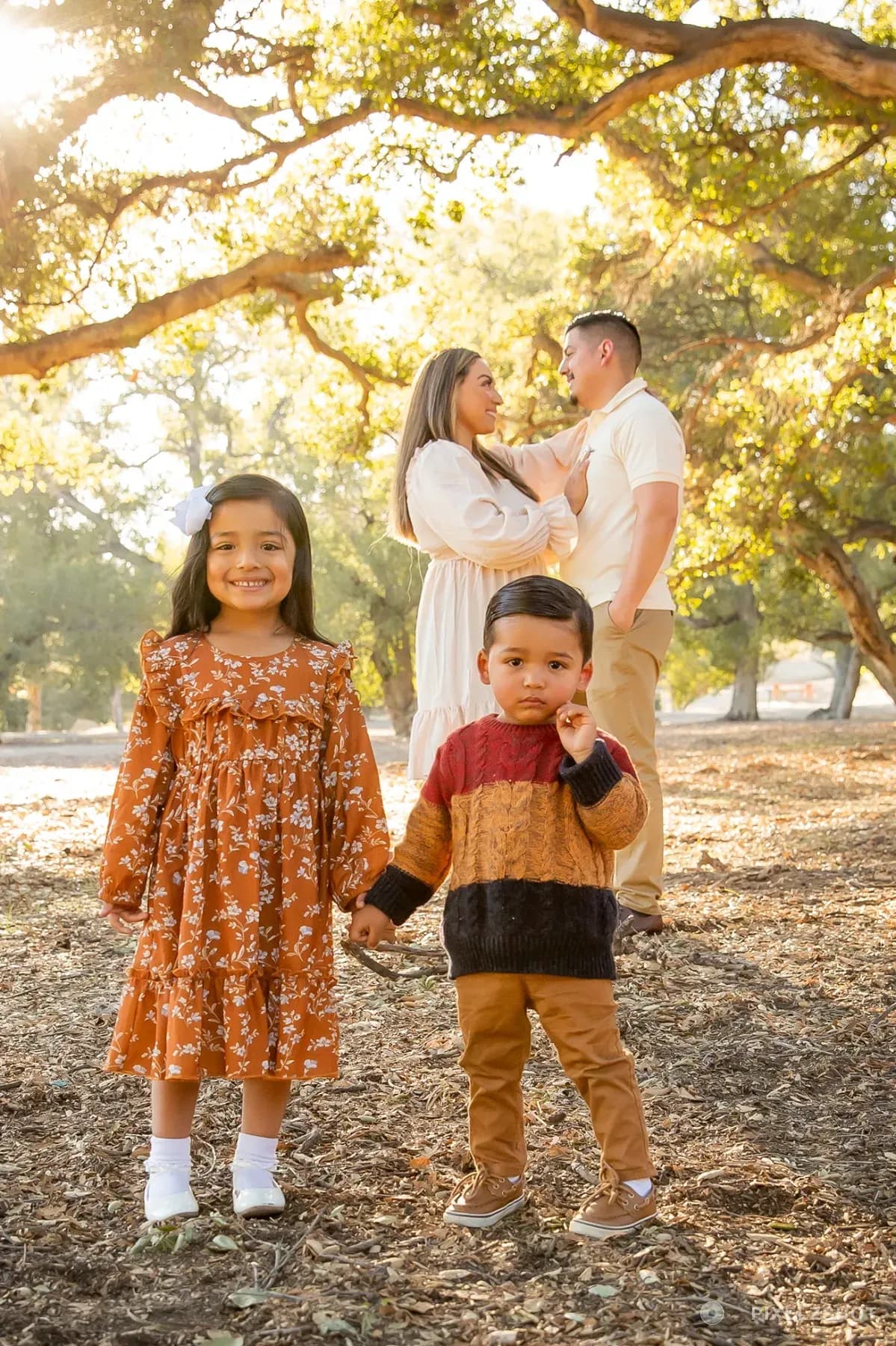 Both children holding hands in the foreground while their parents stand together further back under the branches