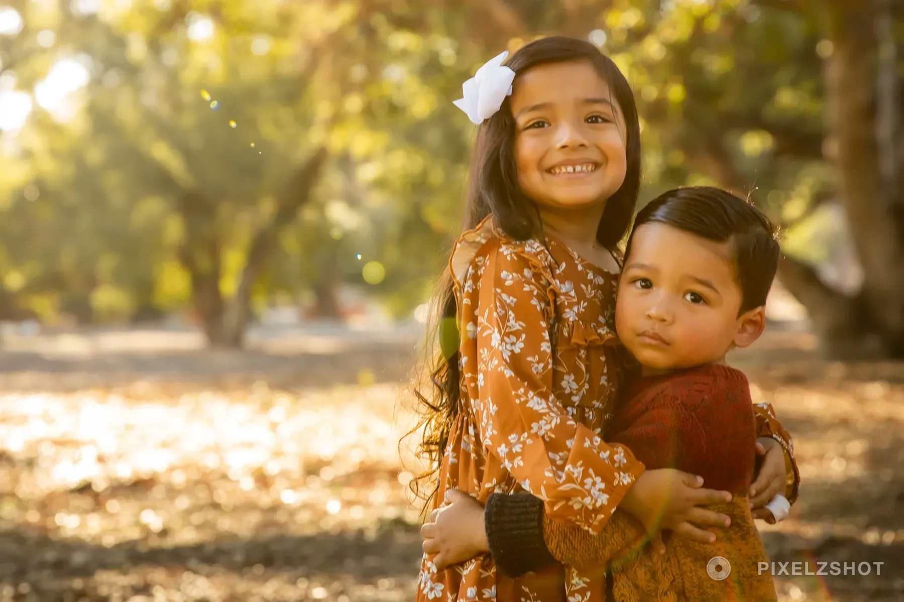 The older sister with both arms around her little brother, grinning at the camera while he looks straight down the lens