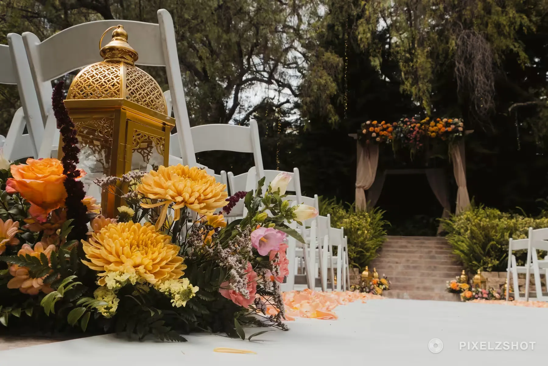 A gold lantern and an arrangement of marigolds and roses at the end of an aisle row, the ceremony arch beyond