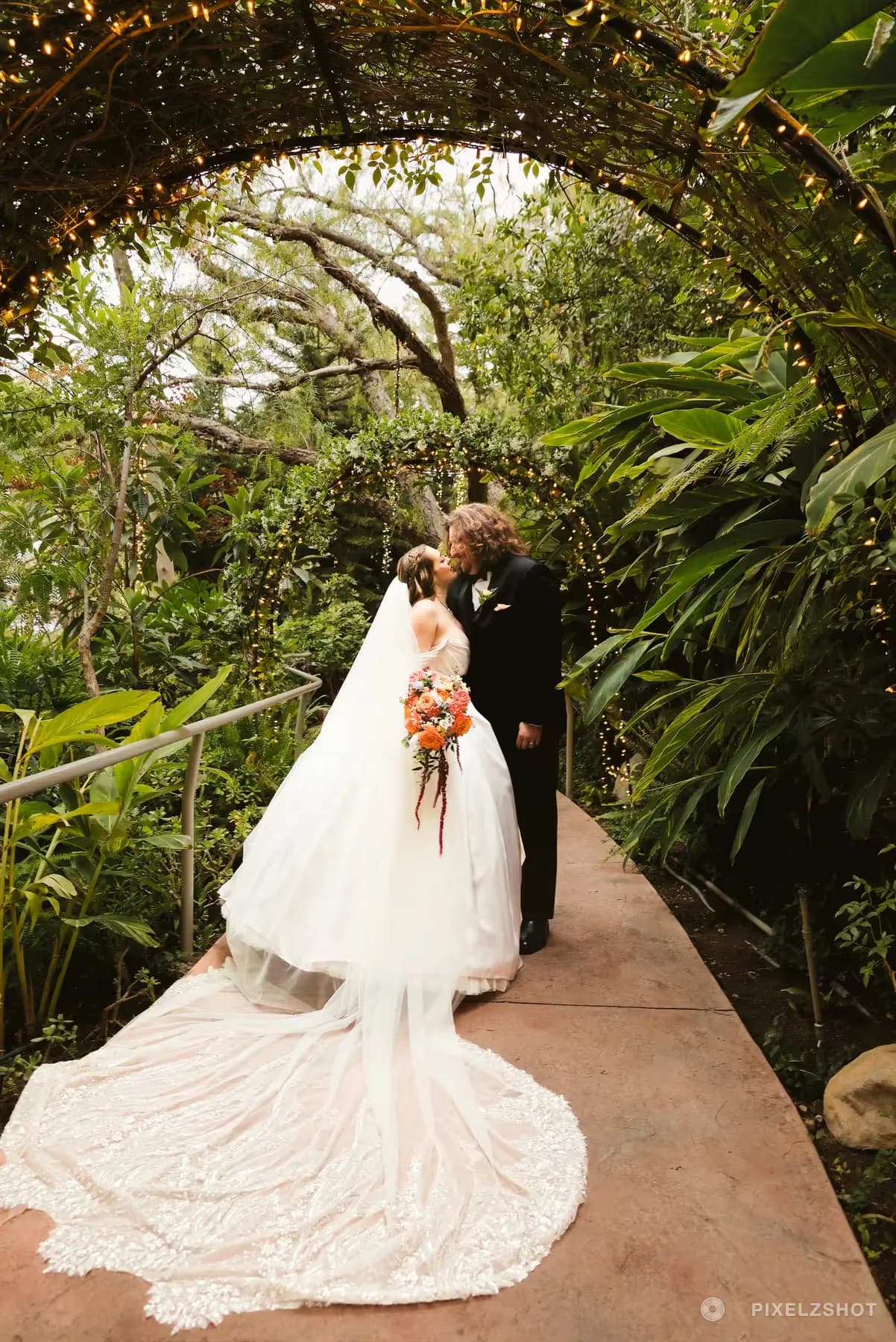 Alex and Adam kissing at the end of a lit garden archway, the lace train spread out along the walkway behind them
