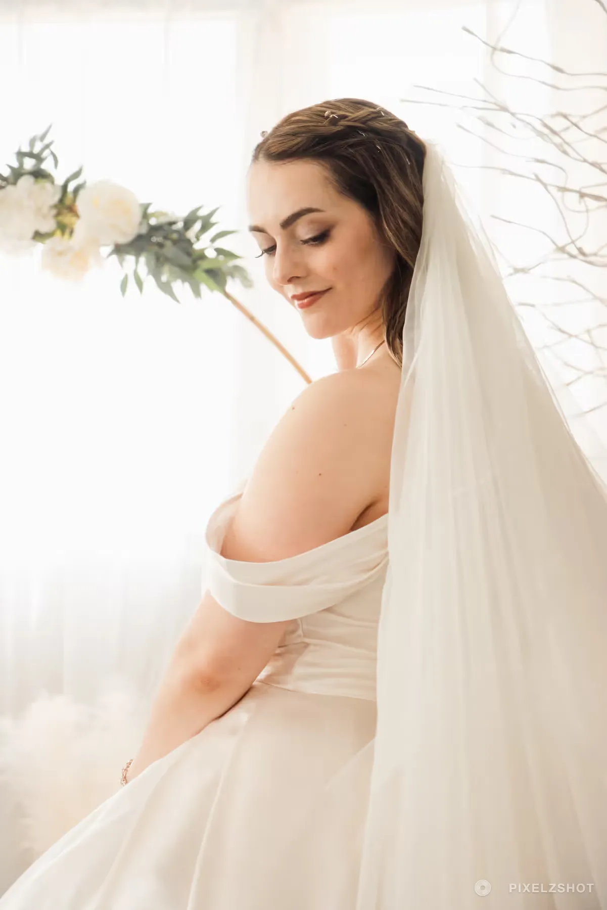 Backlit bridal portrait in an off-shoulder gown with a full-length veil, looking down