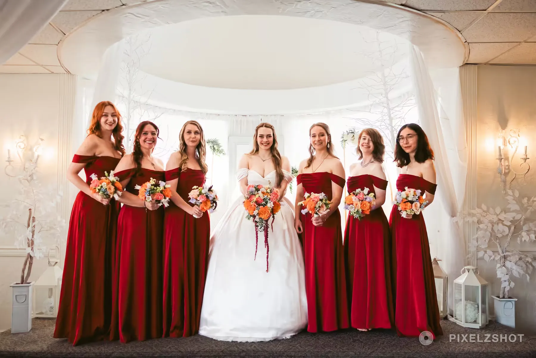 Six bridesmaids in red velvet gowns holding orange and cream bouquets, the bride in the middle under white drapery