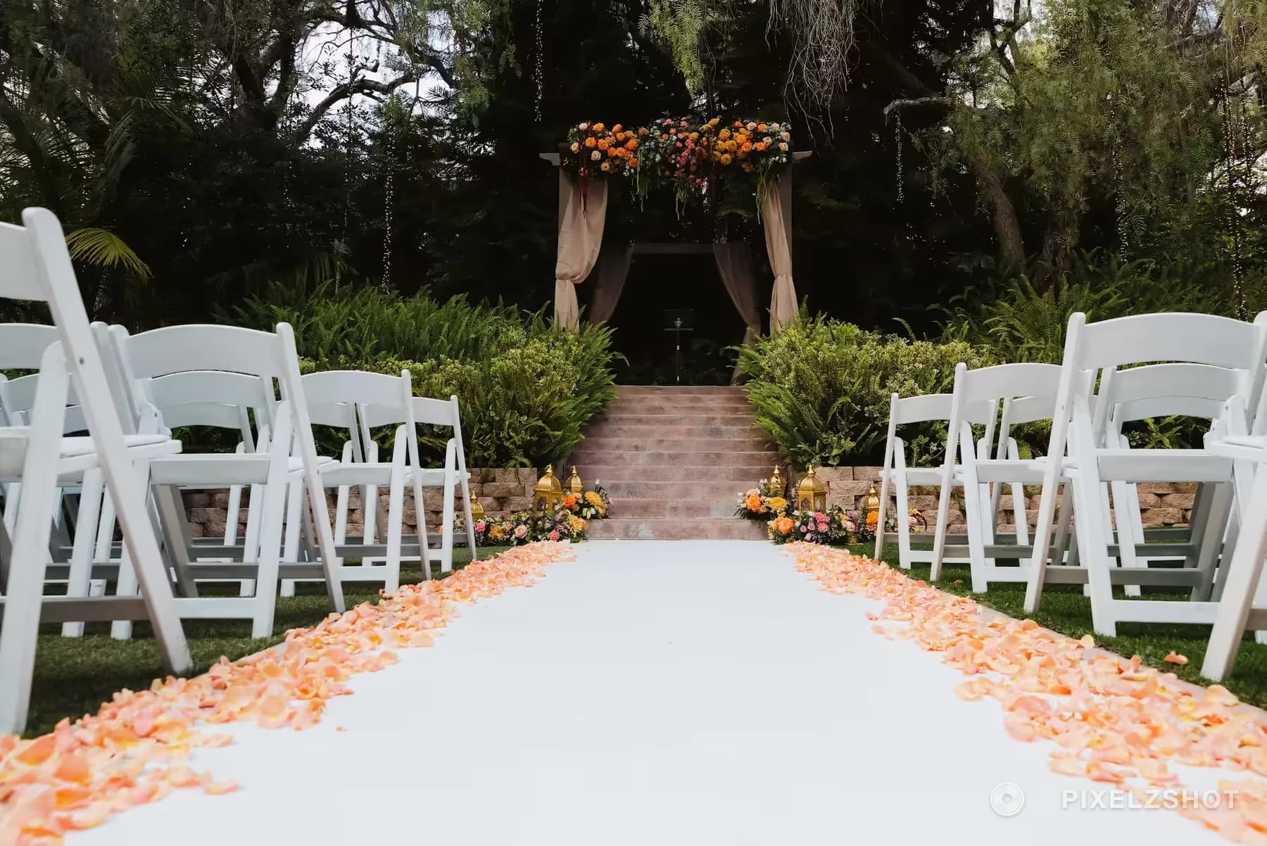The ceremony aisle before guests arrive, a white runner edged with peach petals leading up steps to a draped floral arch