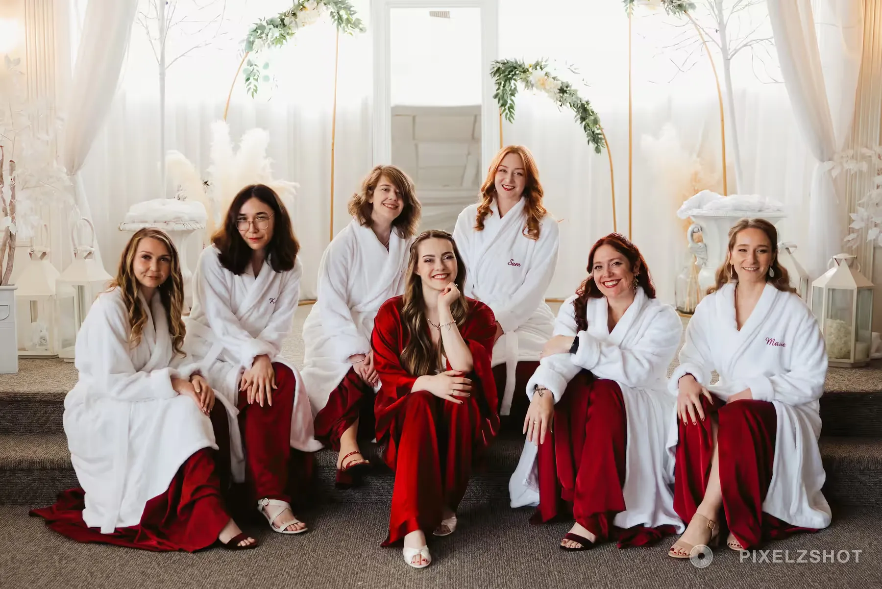 The bridal party seated together in white robes with the bride in deep red, in a suite dressed with pampas and white lanterns