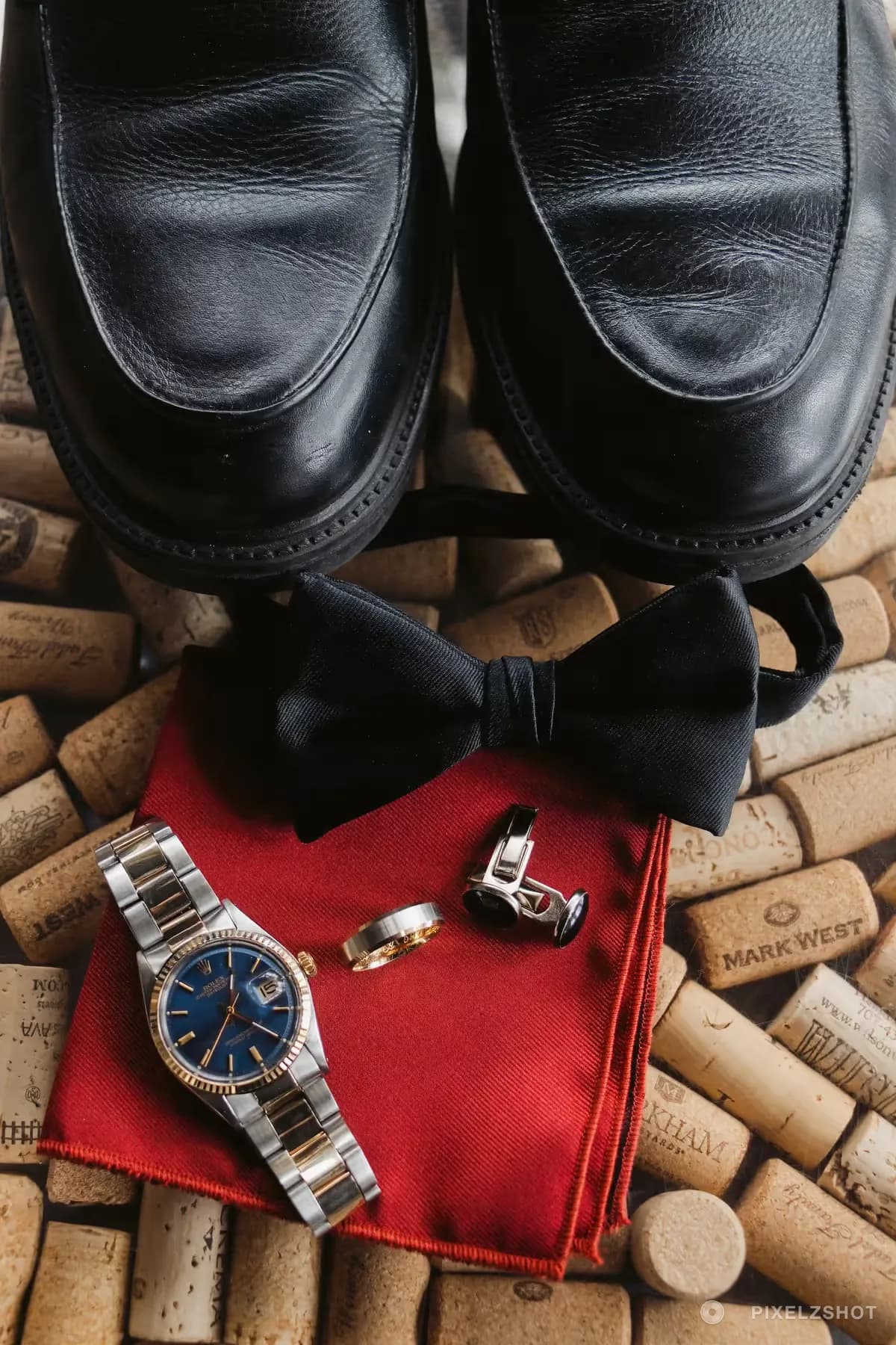 Black shoes, a bow tie, a watch, cufflinks and a wedding band laid out on a red pocket square over wine corks