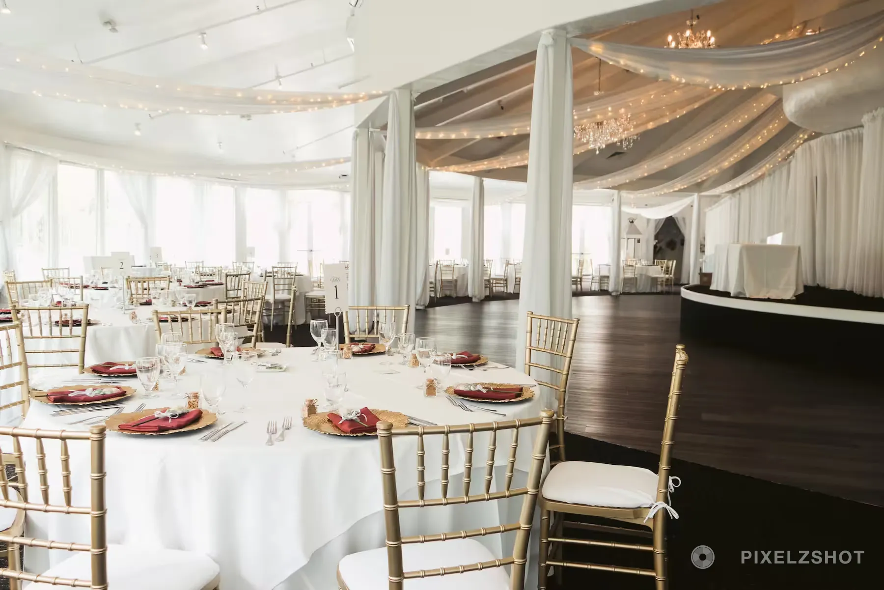 The reception room set with round tables, gold chiavari chairs and red napkins under a draped ceiling of string lights