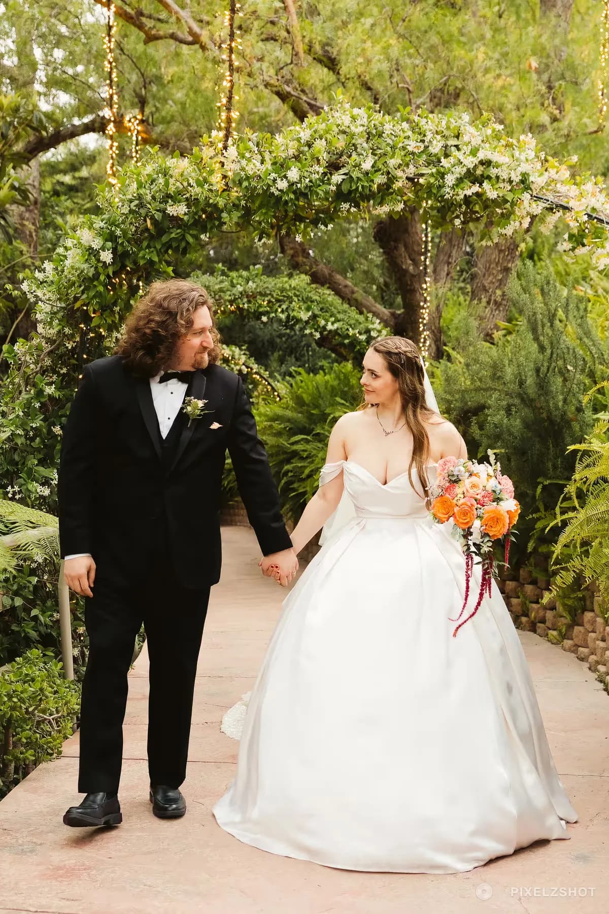 Alex and Adam walking hand in hand along a garden path beneath a flowering trellis strung with lights