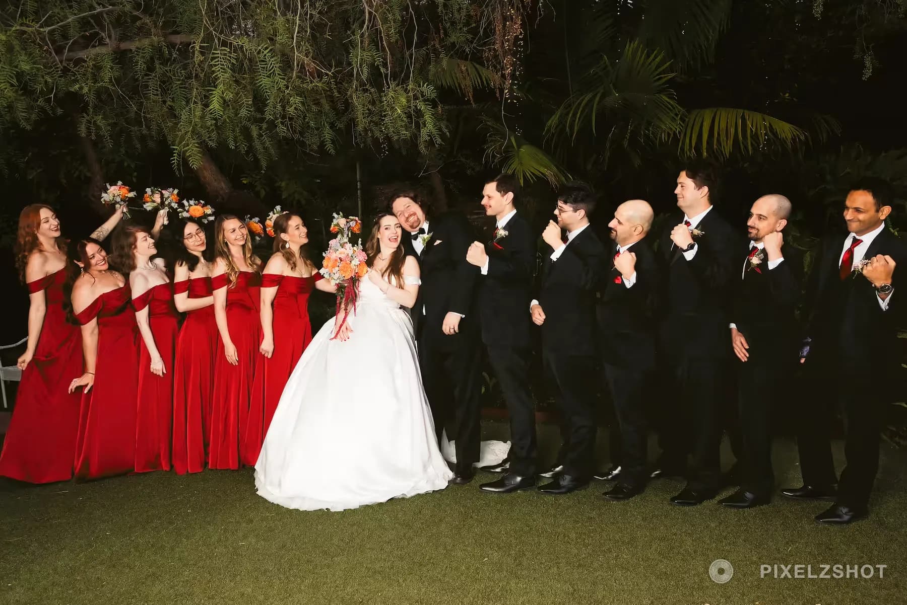 The full wedding party cheering on the lawn at night, bridesmaids in red velvet and groomsmen in black tuxedos