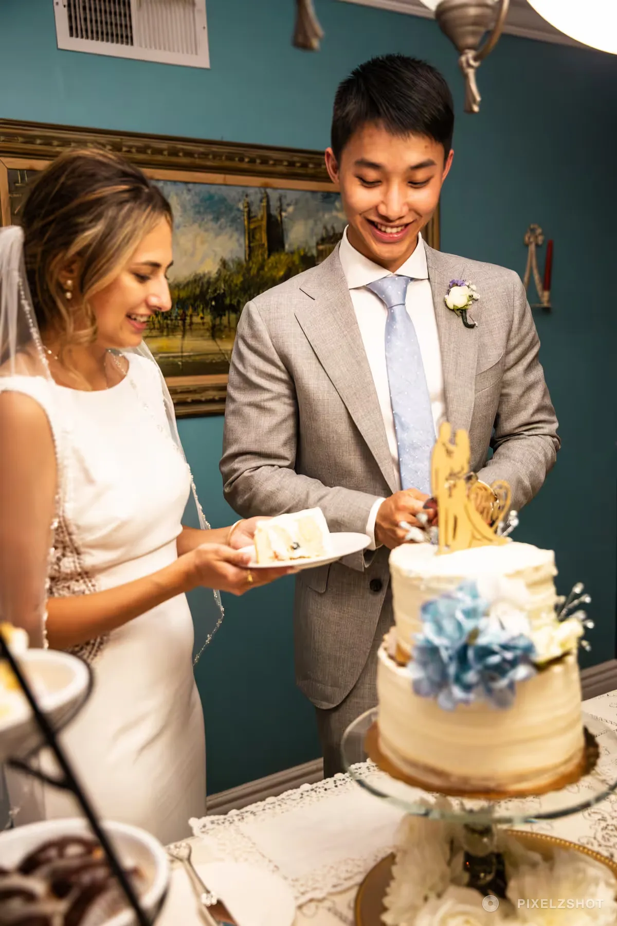 Caleb and Emily cutting a two-tier buttercream cake trimmed with blue hydrangeas, against a deep teal wall