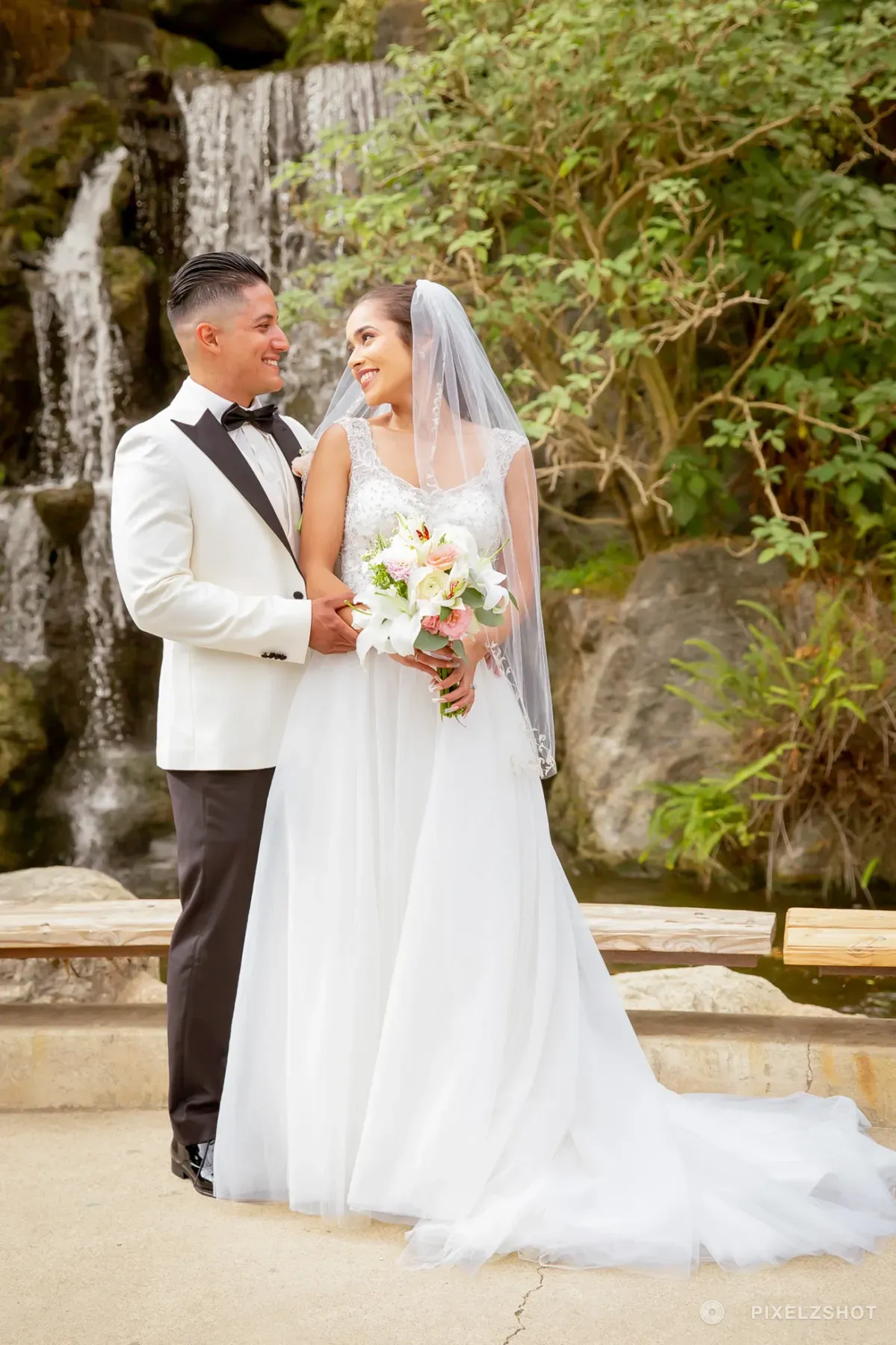 Leslie and Tito holding each other in front of a garden waterfall, her train spread across the stone