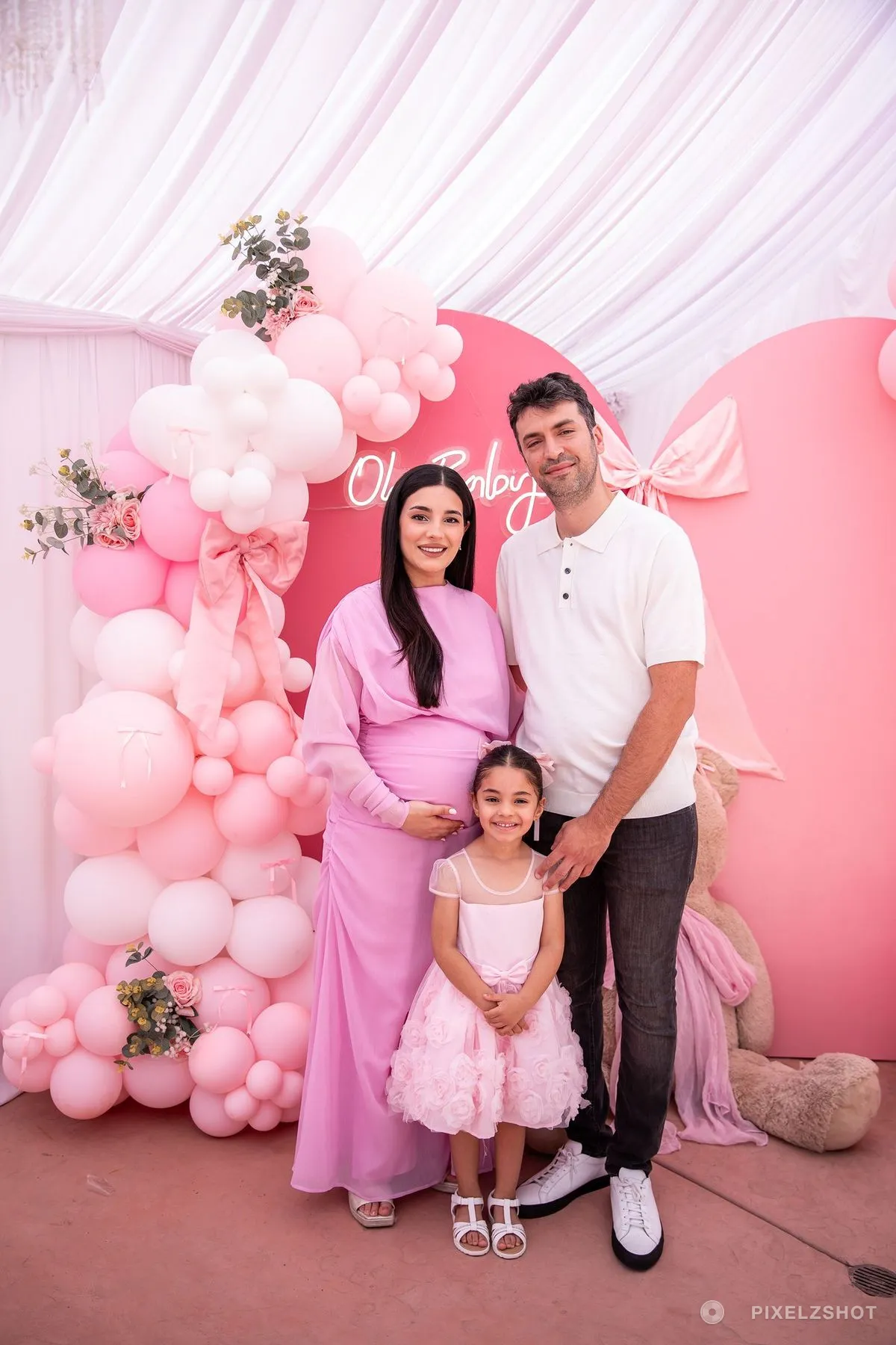 The guest of honour with her family at the pink backdrop, a young girl in a rose dress standing between them