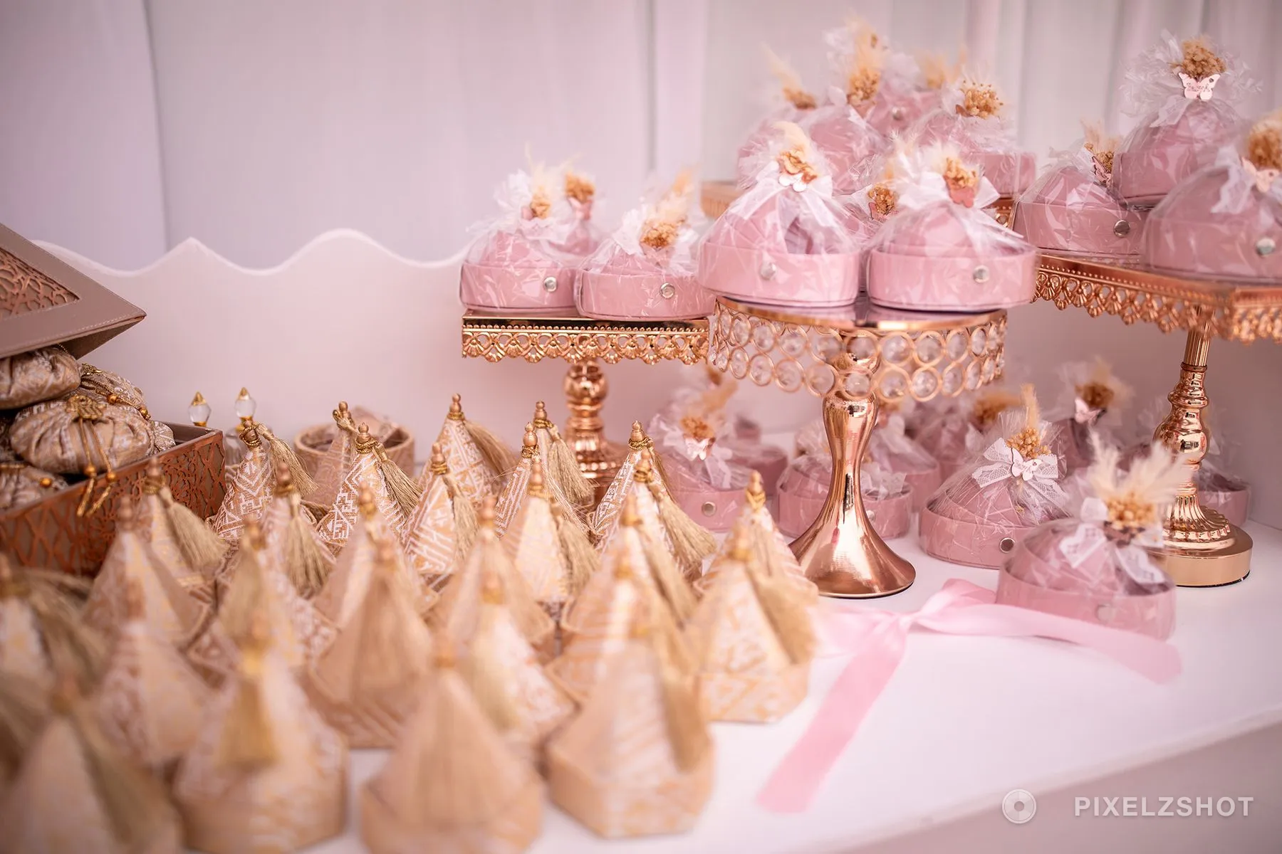 Favor boxes wrapped in cellophane with dried flowers on top, stacked on rose gold stands beside rows of tasselled cones