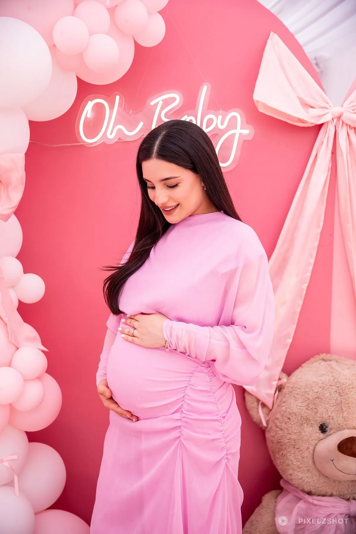 The guest of honour in a pink dress with both hands under the bump, looking down, the Oh Baby neon lit behind her
