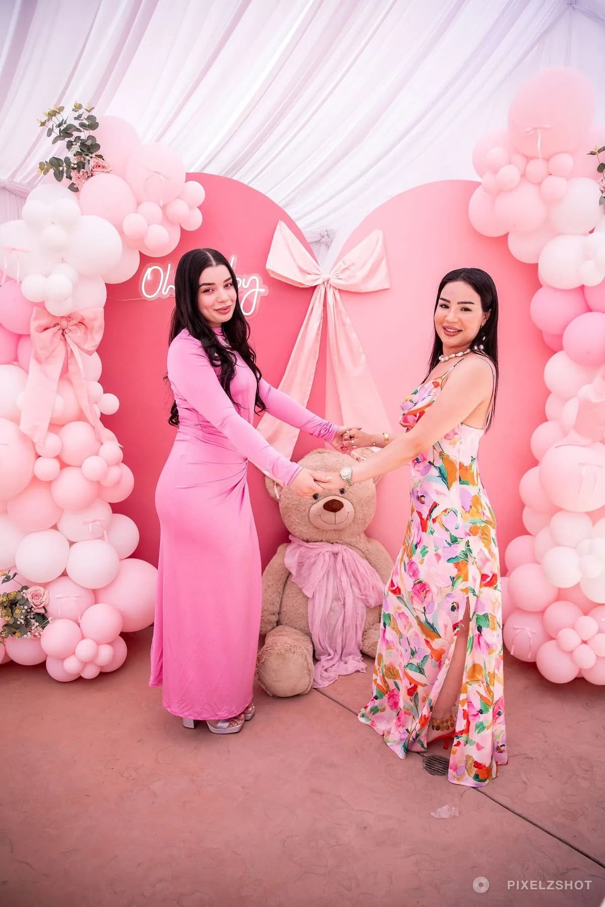 Two guests holding hands and turning to the camera on either side of the giant teddy bear at the backdrop