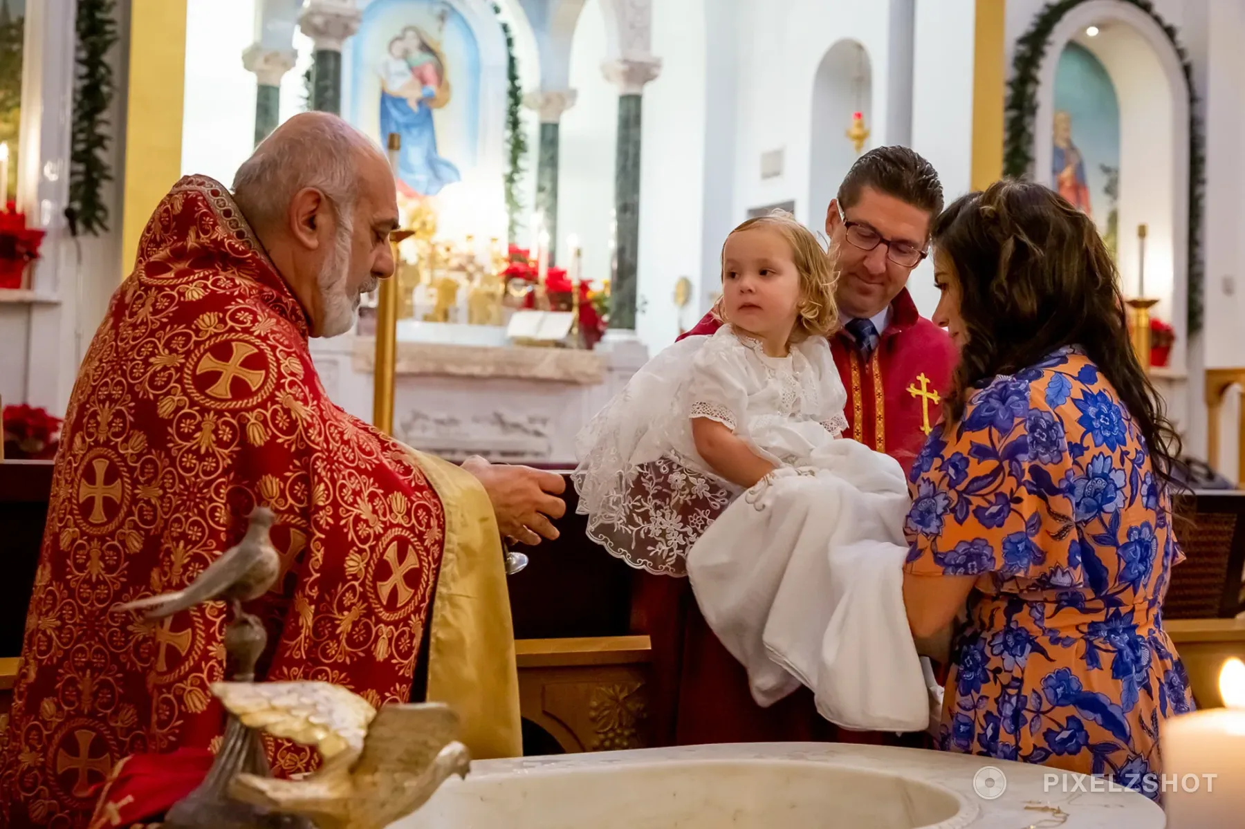 Sierra in a white lace christening gown, held at the font while the priest in red and gold vestments reaches across
