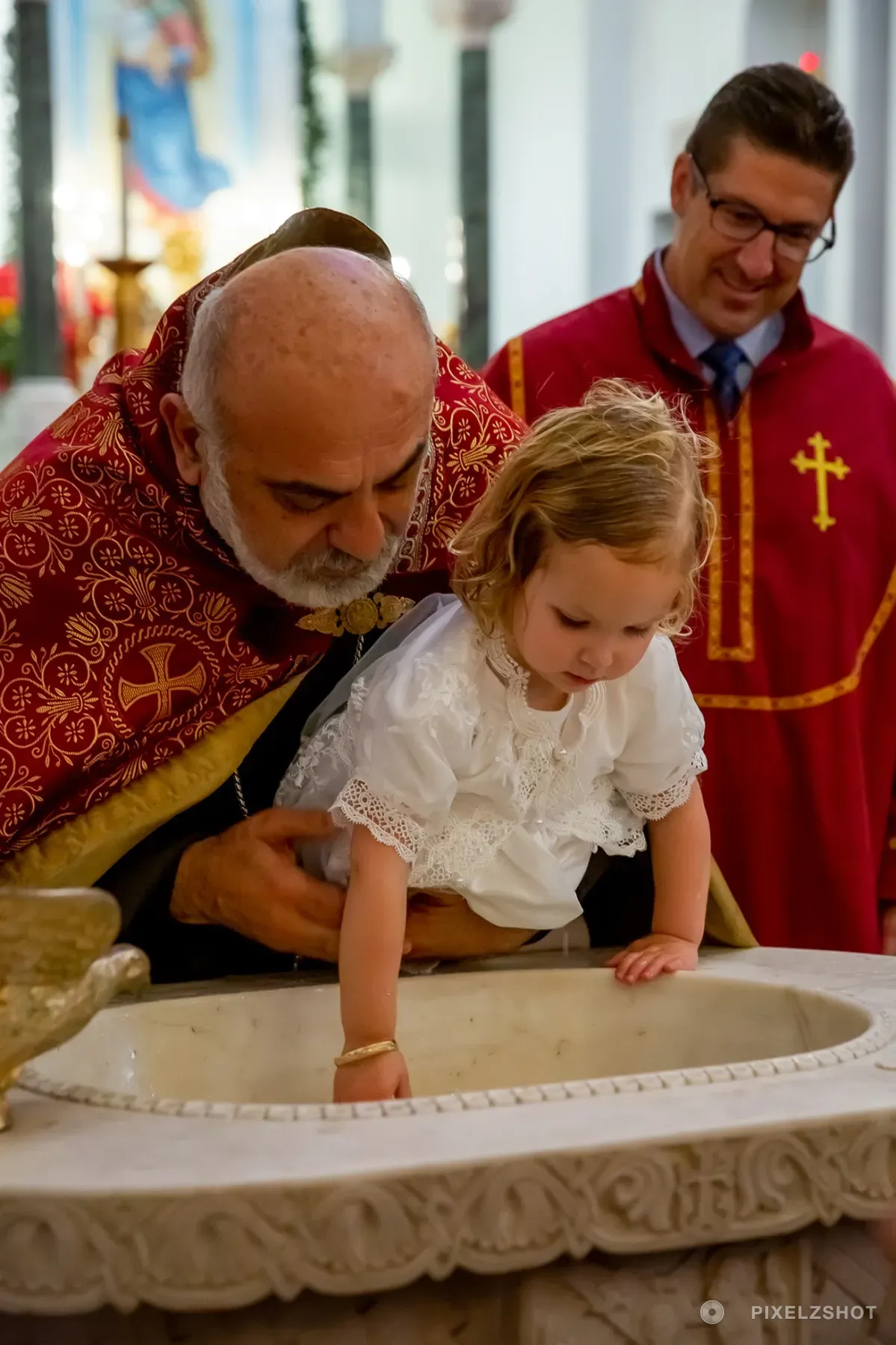 Sierra leaning over the carved marble font with one hand down in the water, steadied by the priest