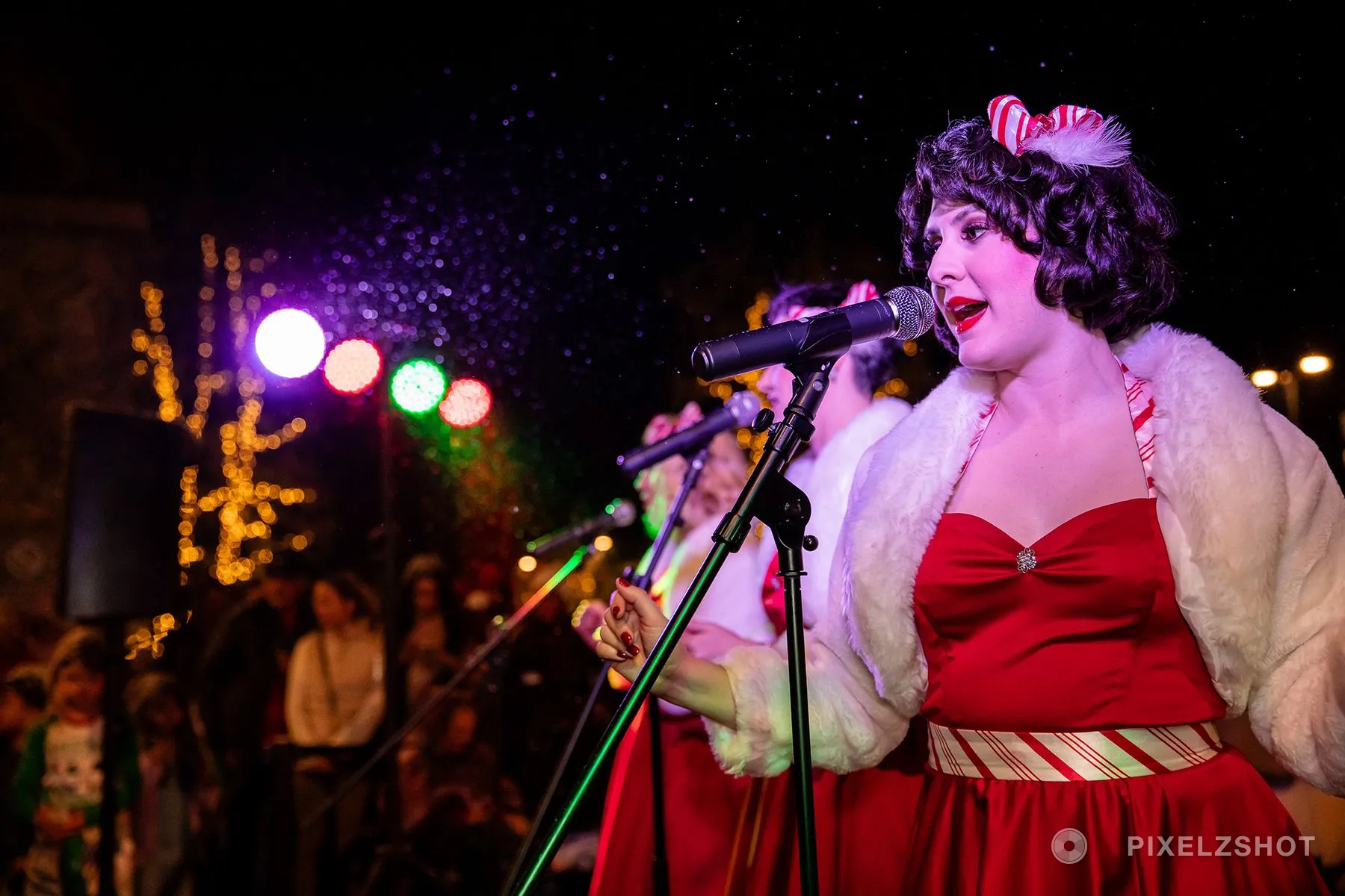 One caroler close at the microphone in a candy-stripe hair bow, coloured stage lights blurred behind her
