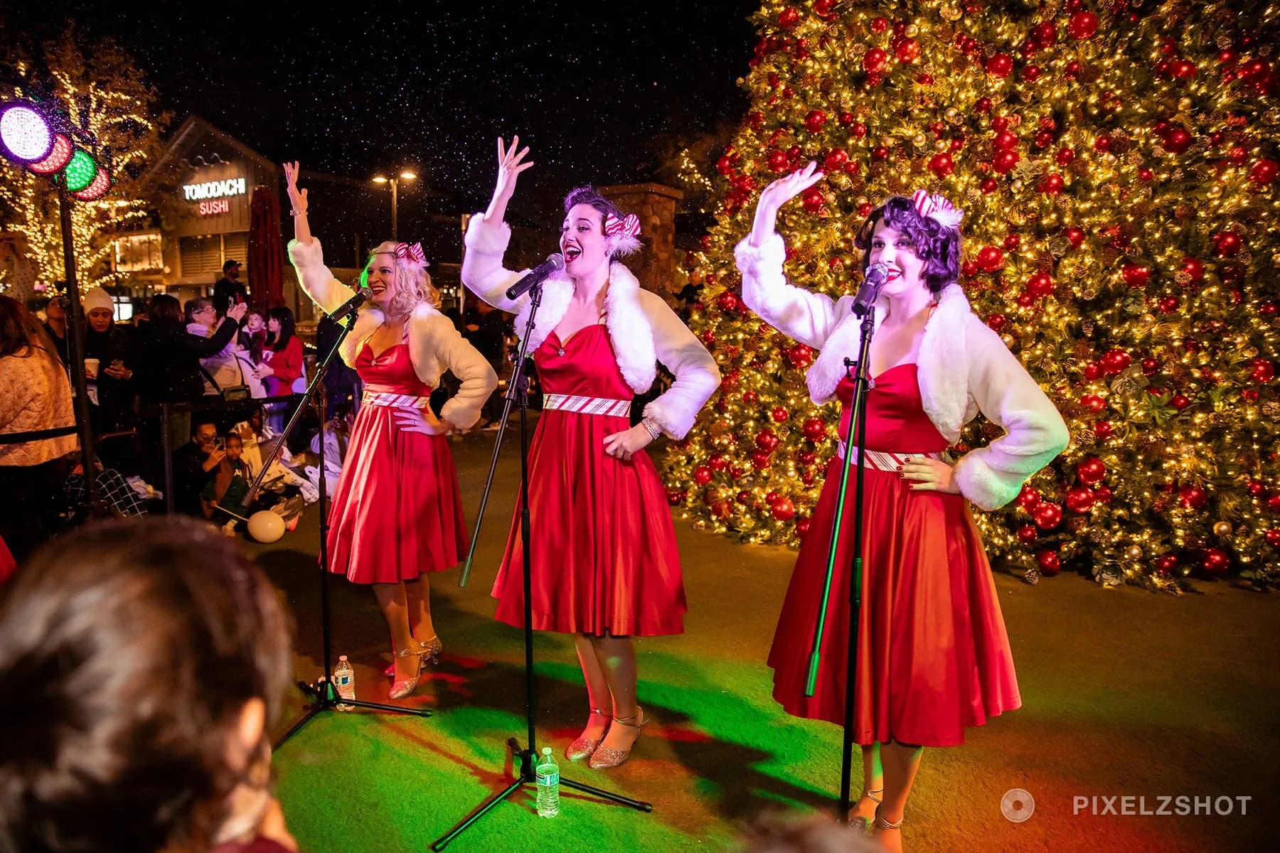 Three carolers in red satin dresses and white fur singing at their microphones beside the lit tree