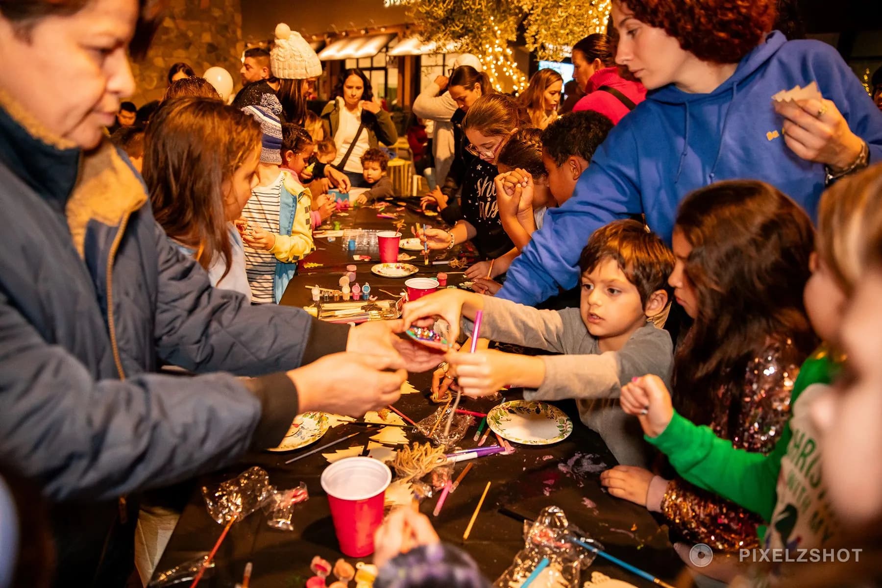 Children crowded along both sides of a long craft table, painting and threading wooden ornaments