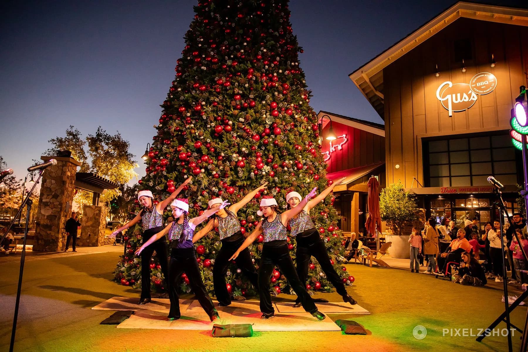 A dance troupe in Santa hats and sequinned tops mid-routine in front of the tree, the sky still blue behind them
