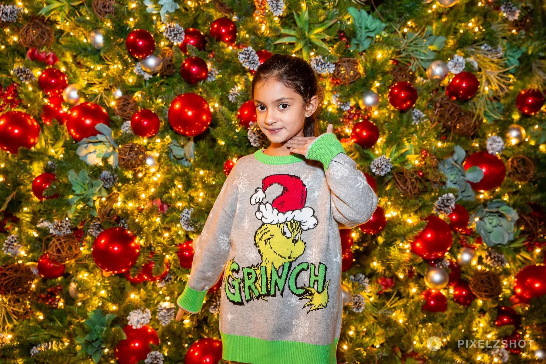 A girl in an oversized Grinch sweater with one hand at her chin, posing against the ornaments