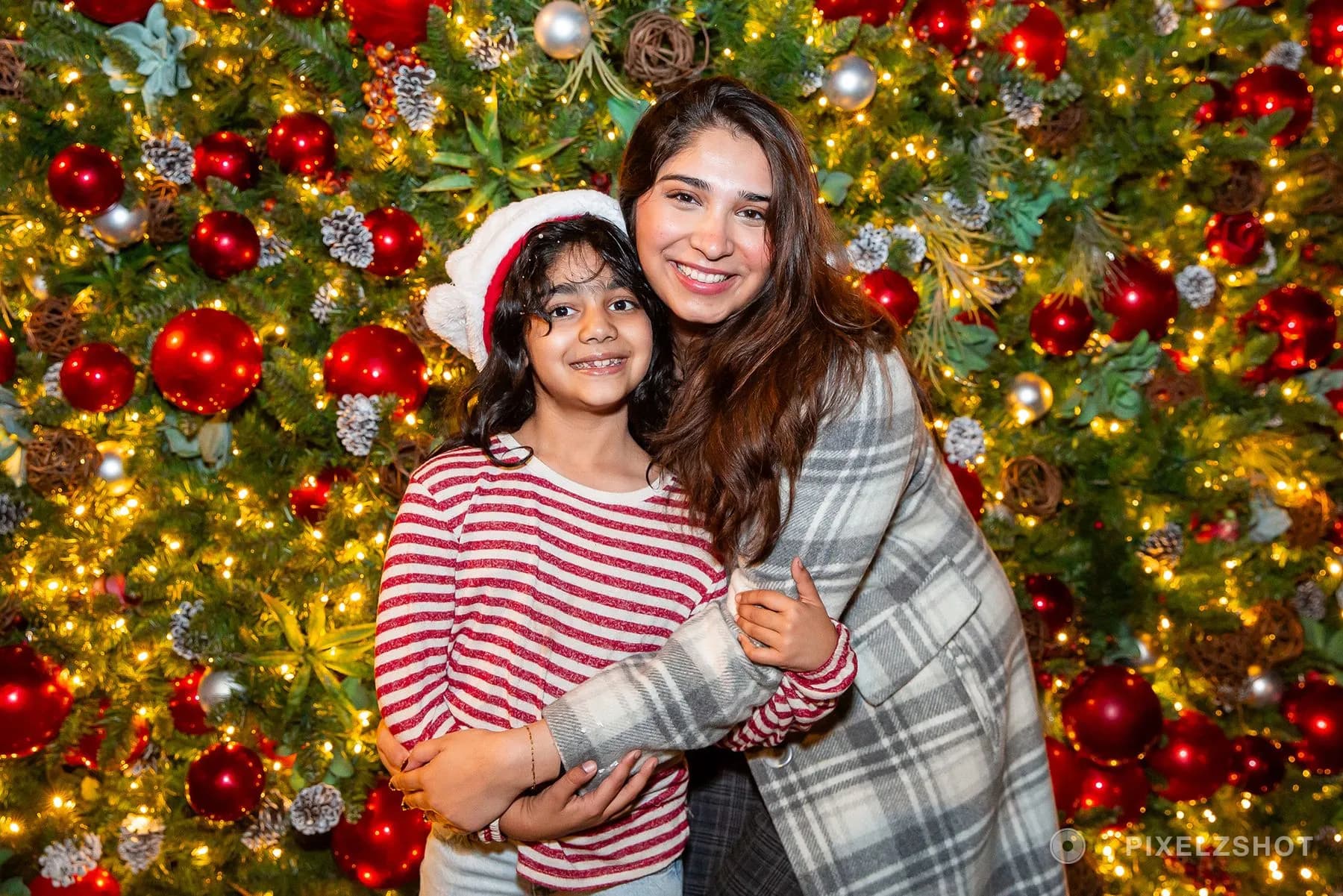 An older girl with her arms wrapped around a younger one in a Santa hat, both smiling at the camera
