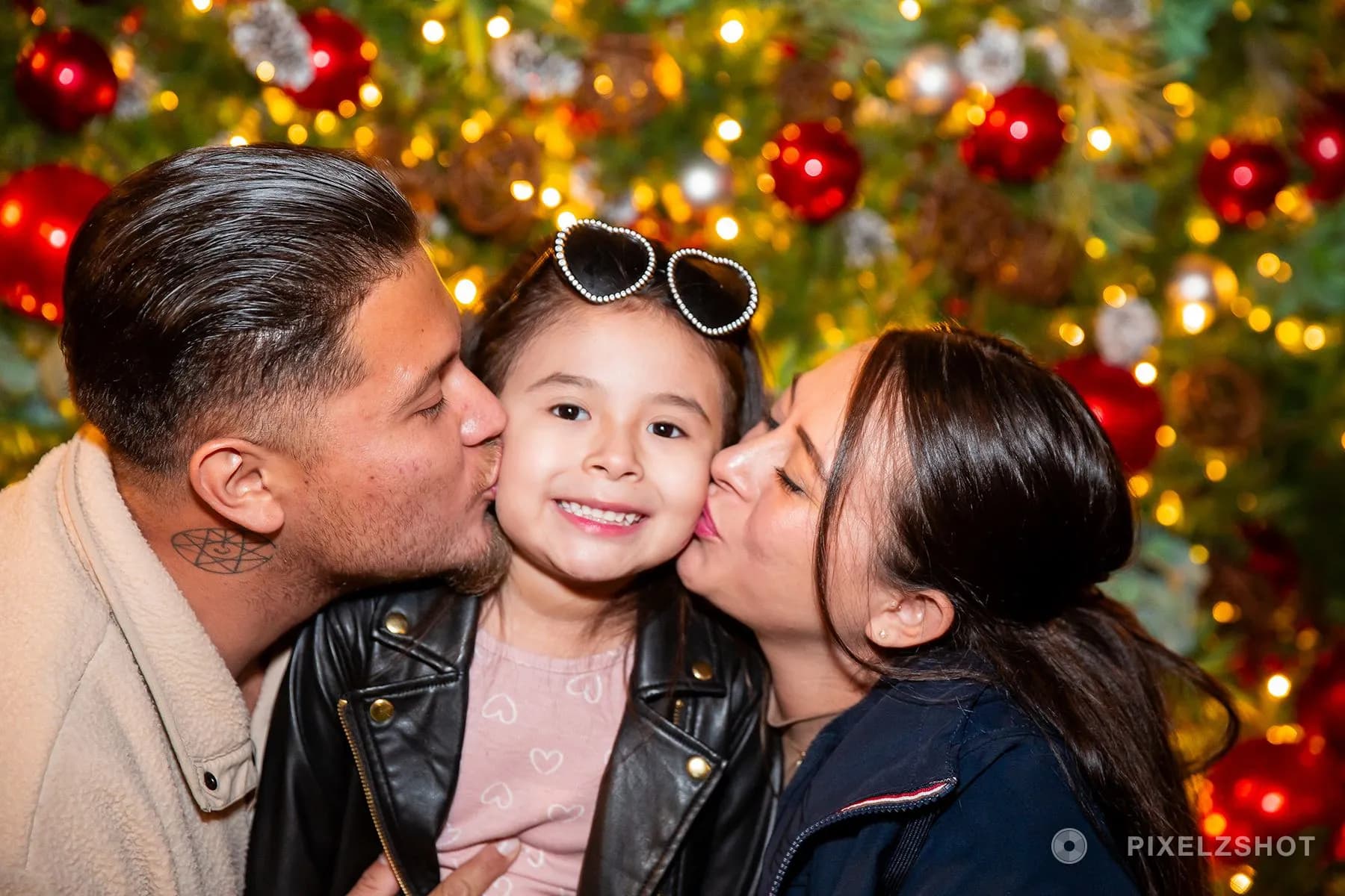 A small girl in heart-shaped sunglasses grinning at the camera while two adults kiss her cheeks at once