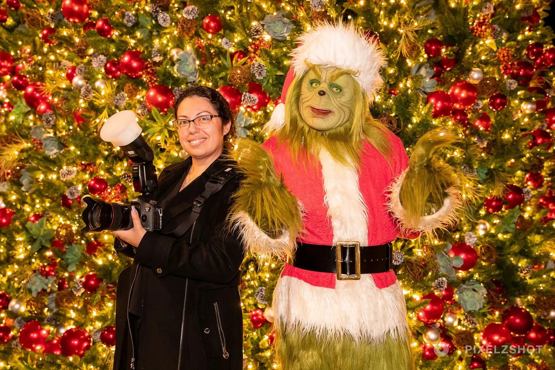 The photographer with her camera and flash standing beside the Grinch in front of the lit tree