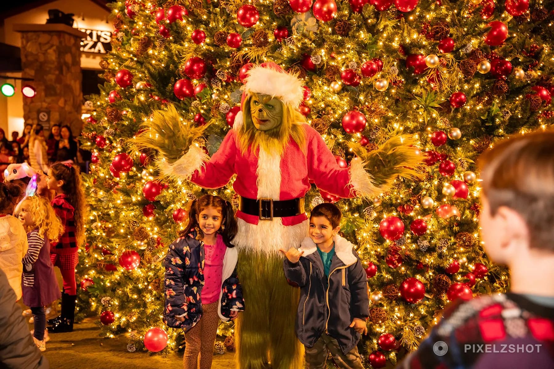 The Grinch in a Santa suit with arms spread wide behind two grinning children in front of the tree