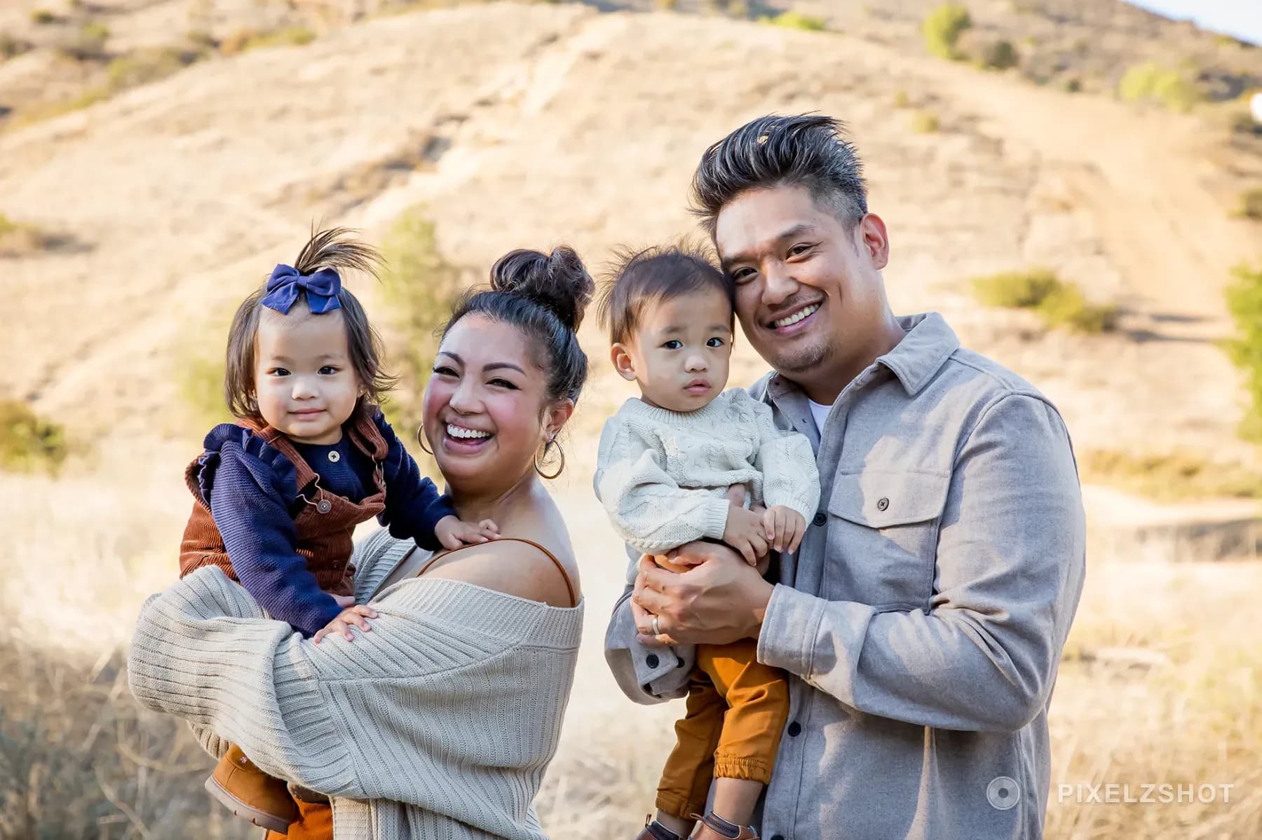 Both parents holding a toddler each, all four laughing at the camera with a gold canyon hillside behind them