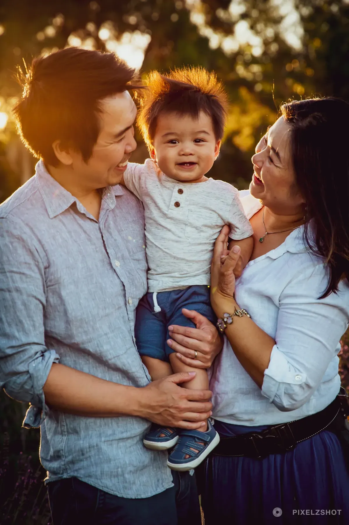 Both parents holding their laughing toddler between them, backlit so the sun catches the top of his hair