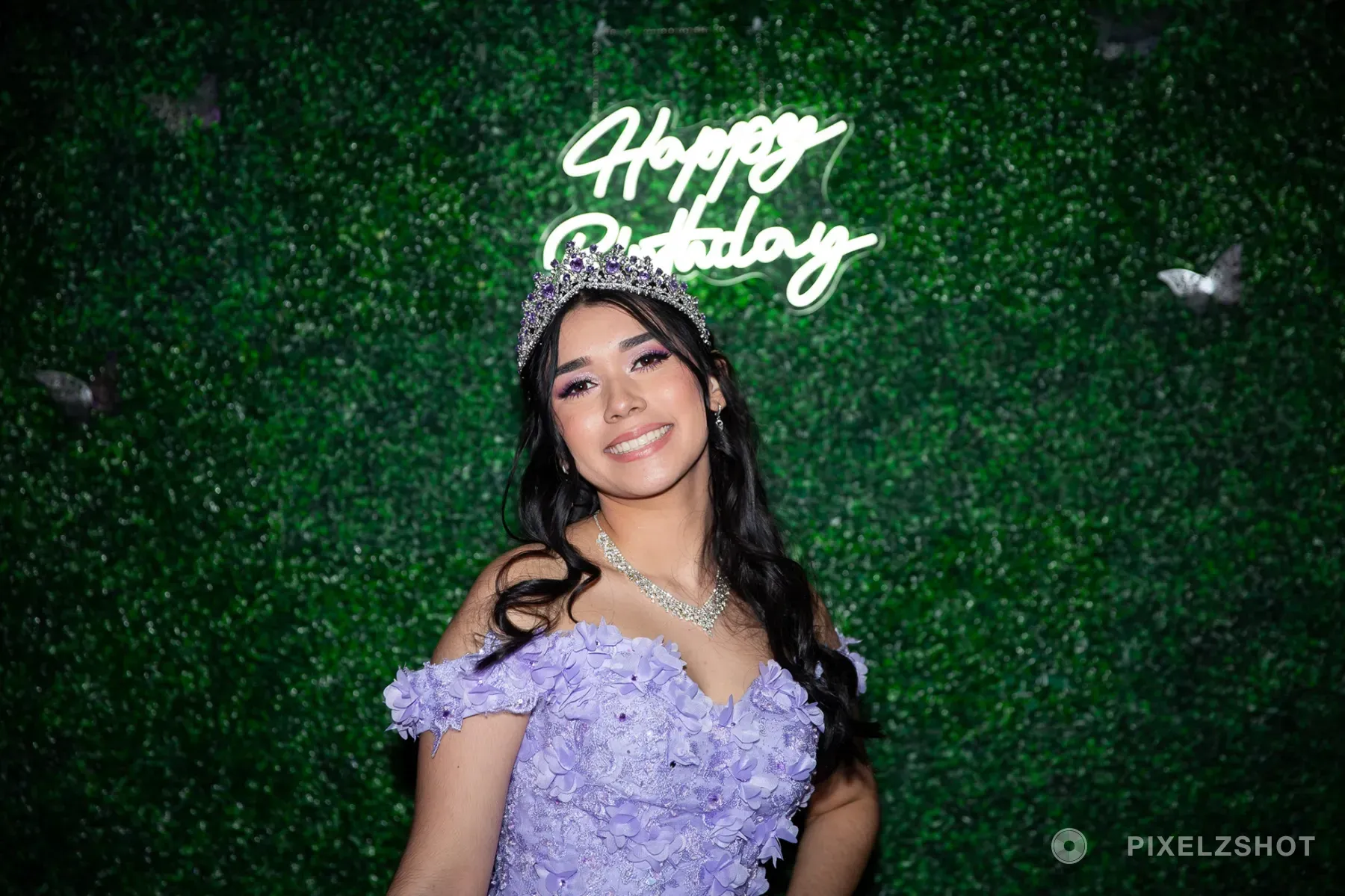 Jasmine in her crown and crystal necklace, grinning at the camera with the Happy Birthday neon glowing behind her