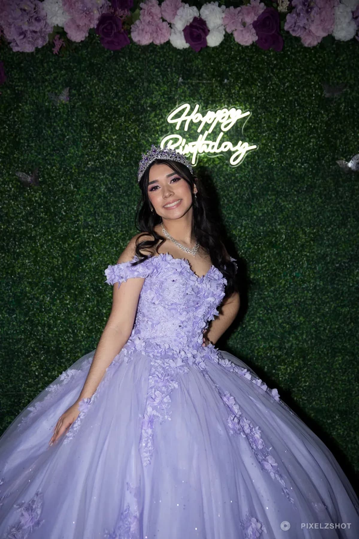 Jasmine full length against a boxwood wall under a Happy Birthday neon sign and a garland of lilac and white roses