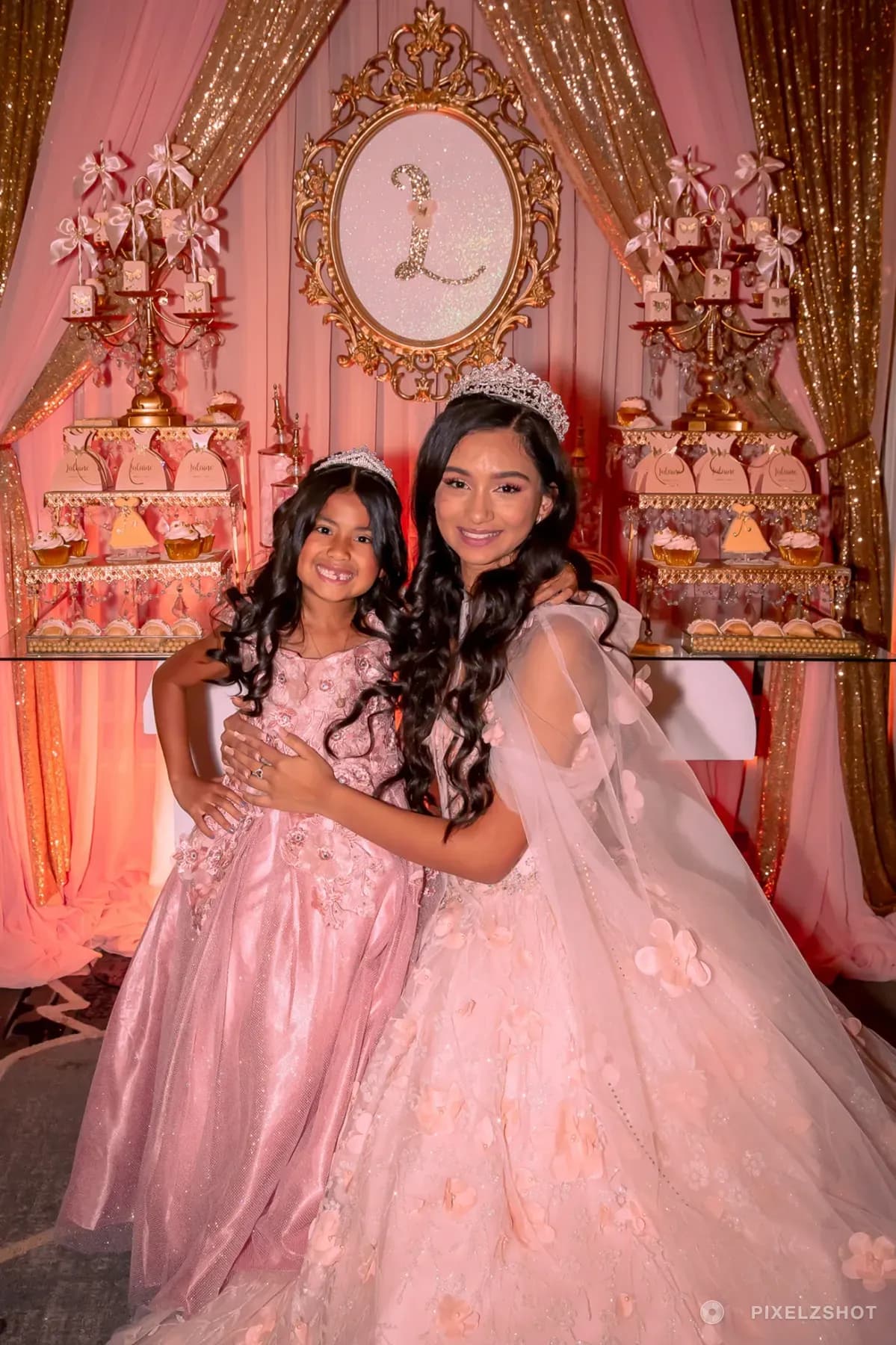 Lalaine holding her younger sister close in front of the blush and gold backdrop, both in matching pink and crowns