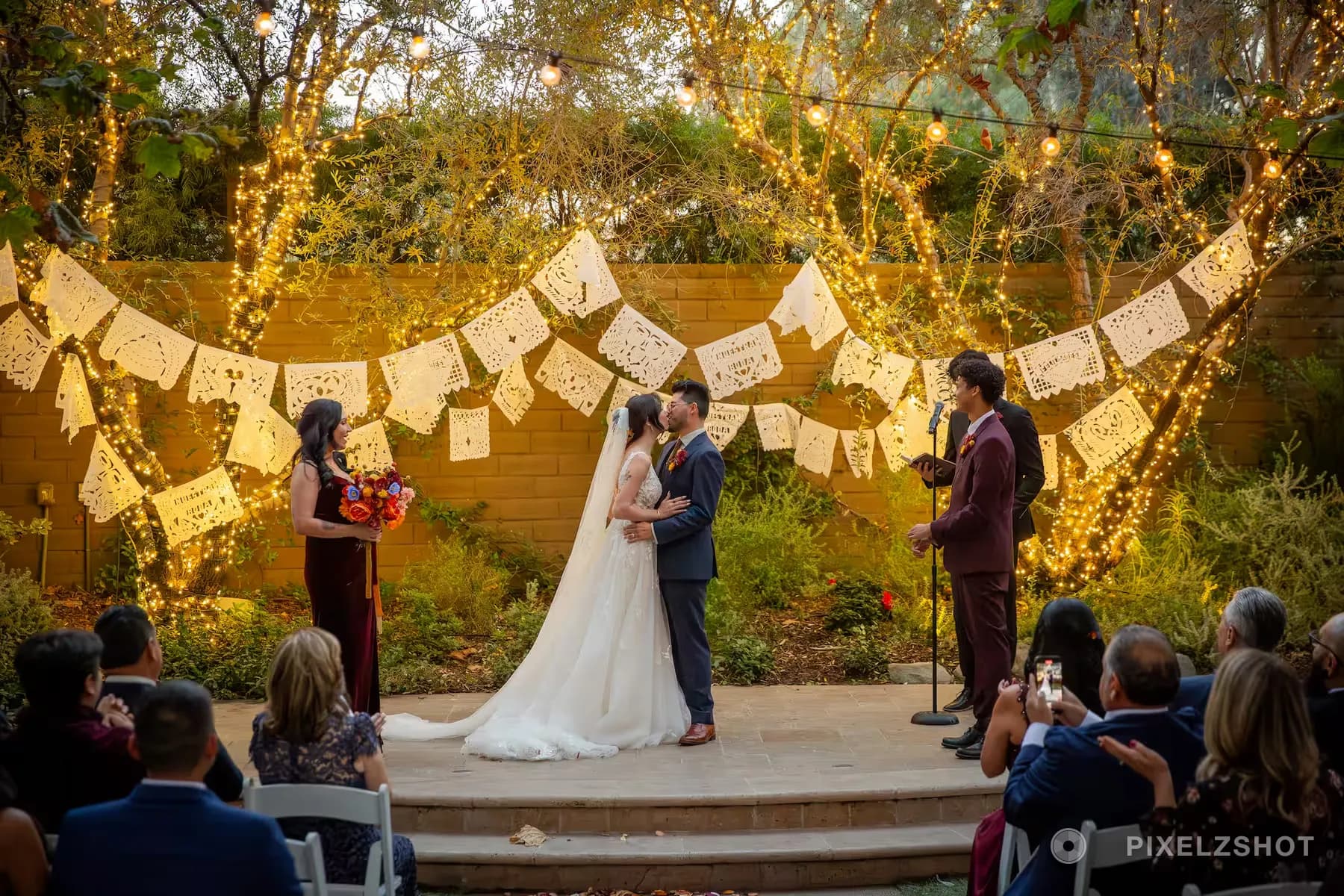 Danielle and Matthew kissing on the ceremony platform beneath rows of papel picado and string lights, guests seated in front