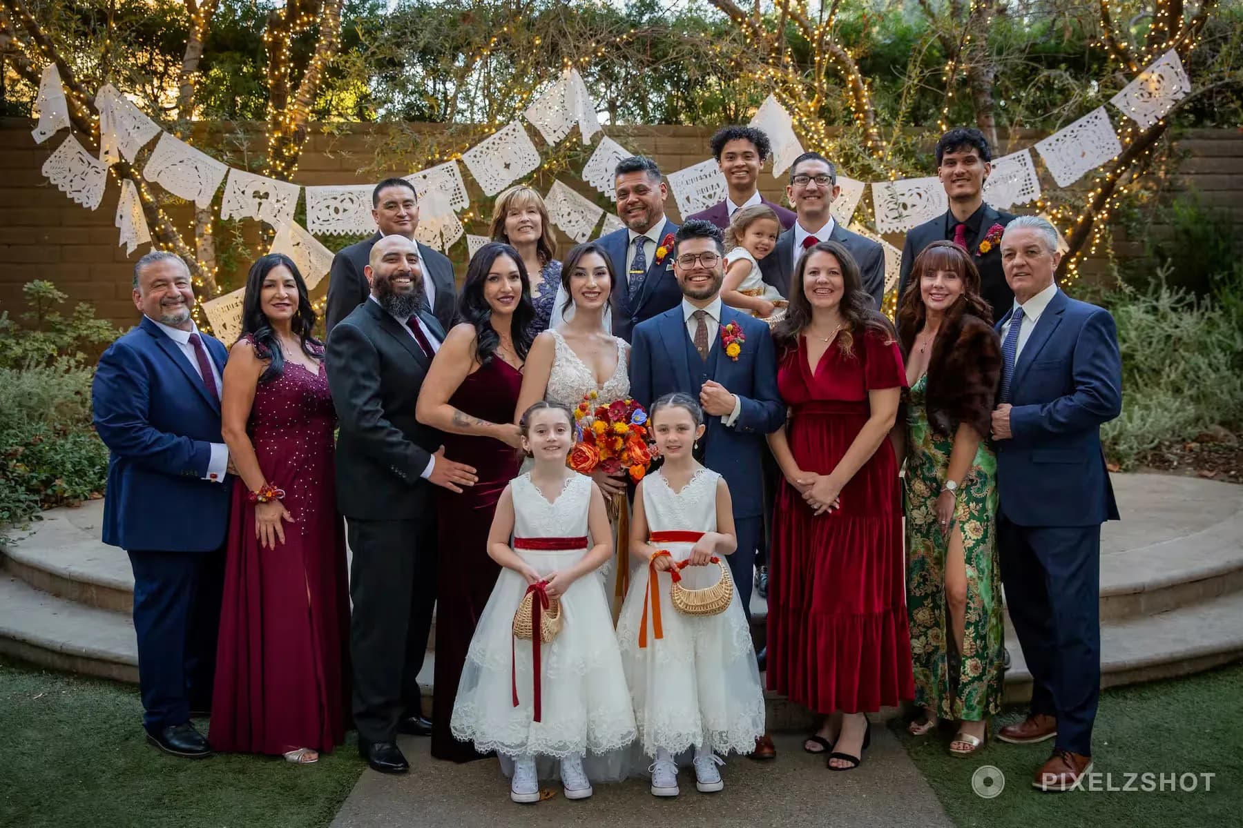 A large family group portrait under papel picado banners and string lights, flower girls with baskets at the front