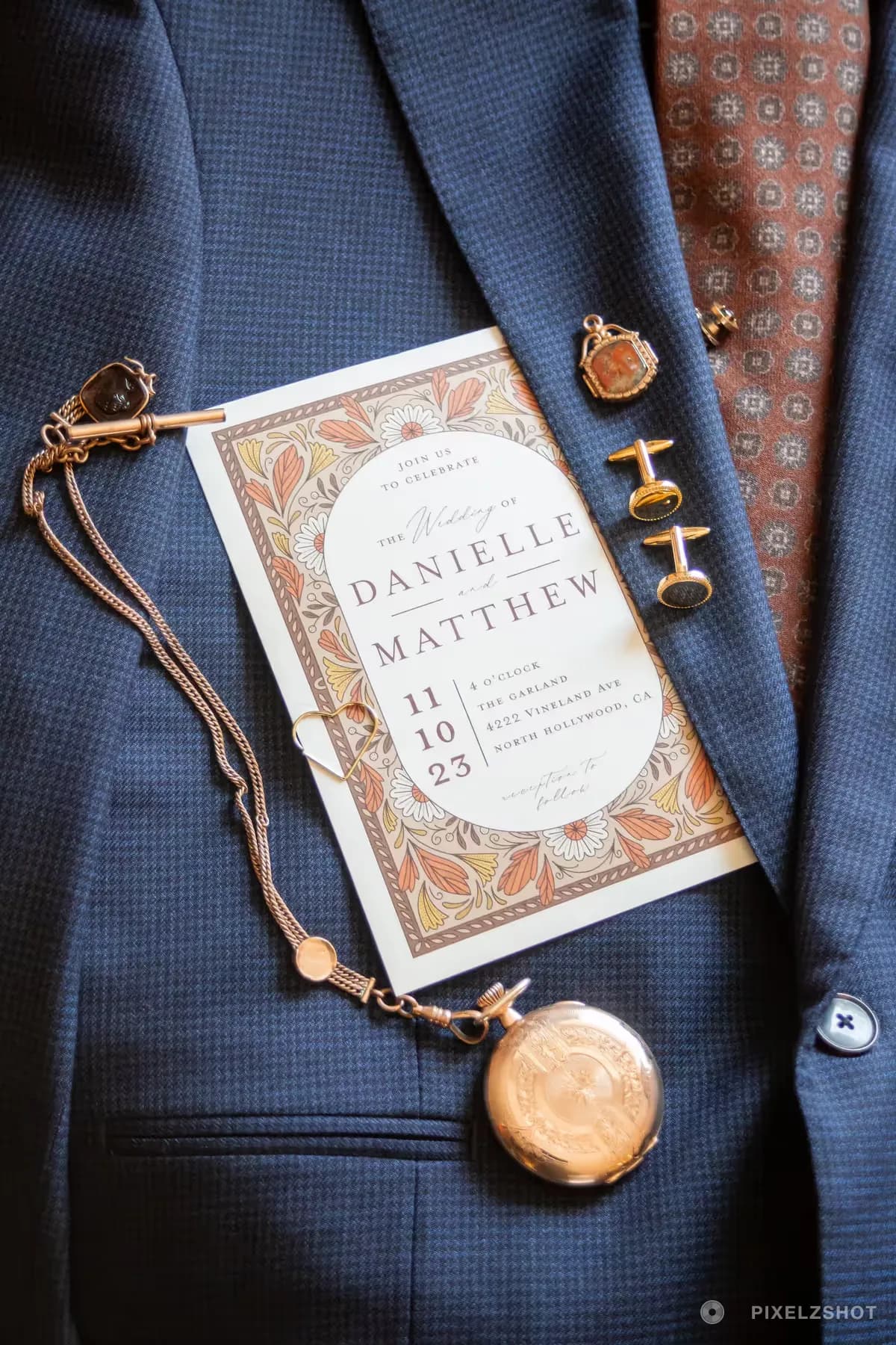 The wedding invitation with a folk-art floral border, laid on a navy jacket beside a pocket watch, cufflinks and a rust-patterned tie