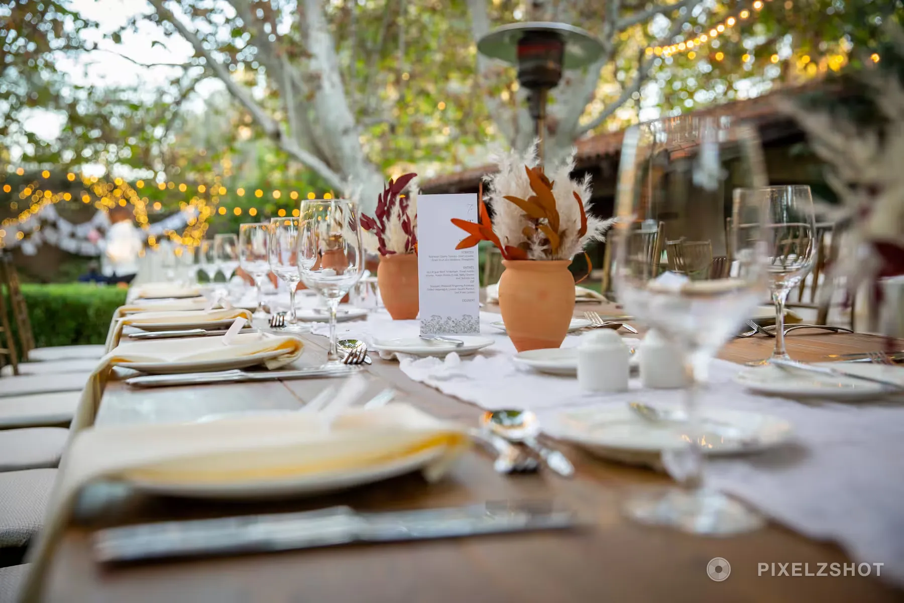 A long reception table under the trees set with terracotta jugs of dried orange florals, menu cards and glassware