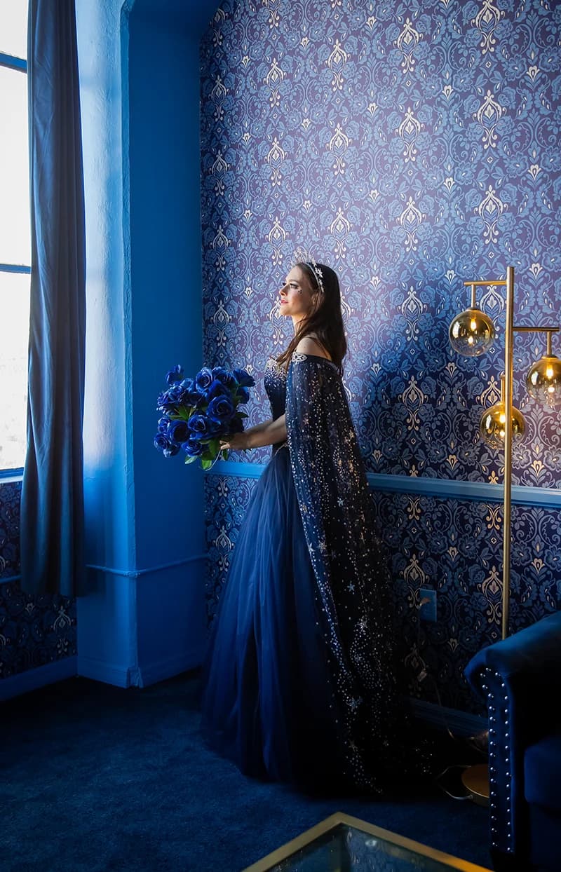 Woman in a starry blue gown and cape holding a bouquet of blue roses by a window