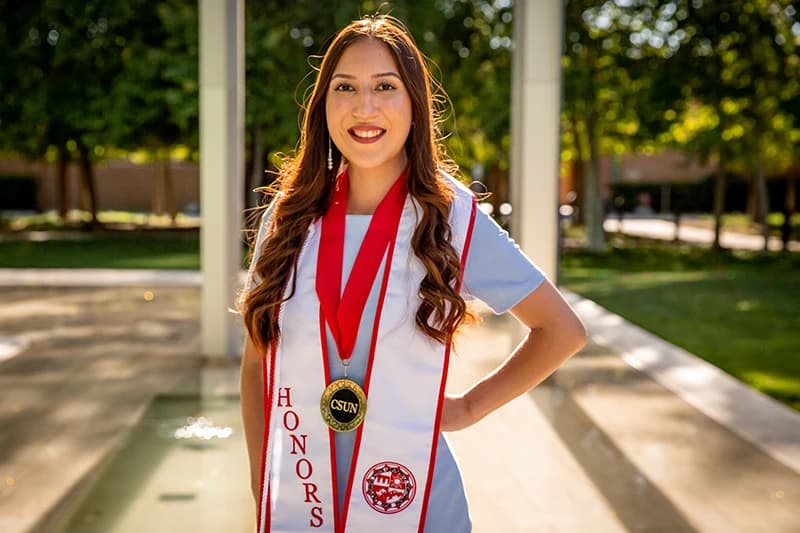 Graduate in an honors stole posing on a tree-lined campus walkway